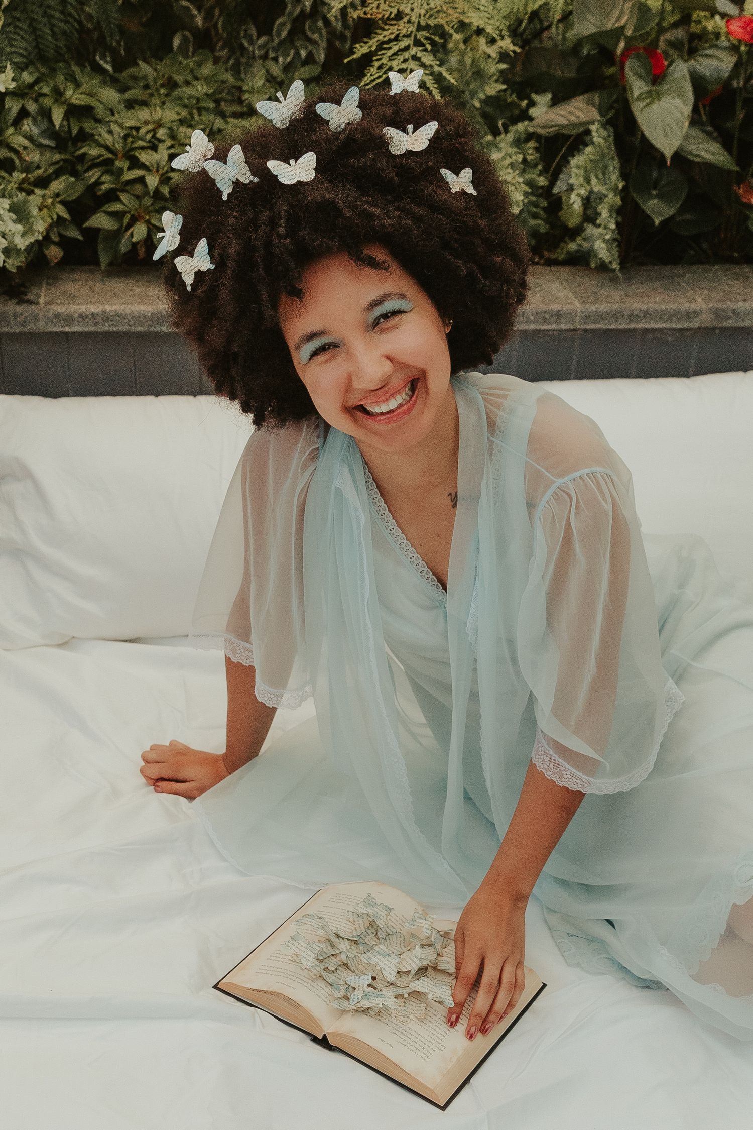 Person in light blue dress lying on white bedding with decorative butterflies and reading a book.