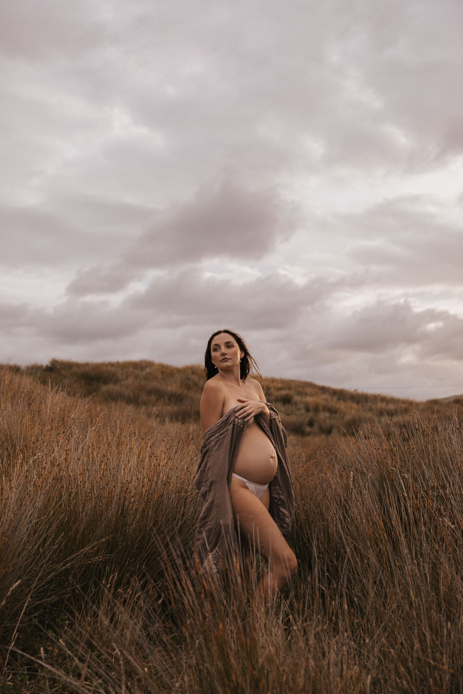 Dramatic maternity portraits in tall grass with overcast sky creating moody atmosphere.