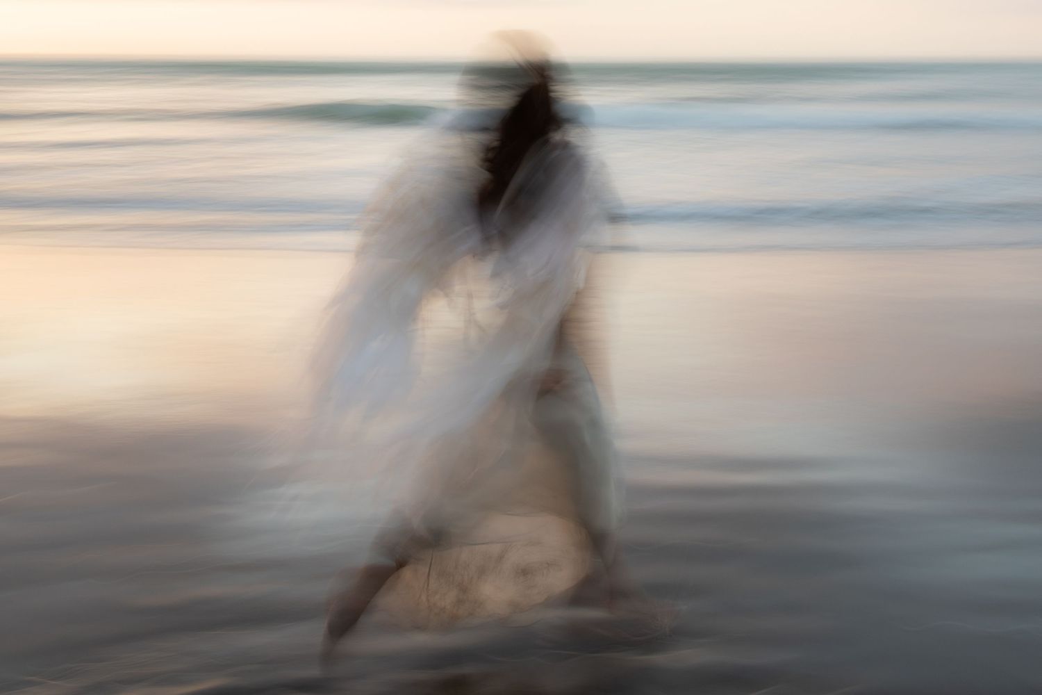 A motion-blurred figure in white walks along a misty beach shoreline at sunset creating an ethereal silhouette effect.