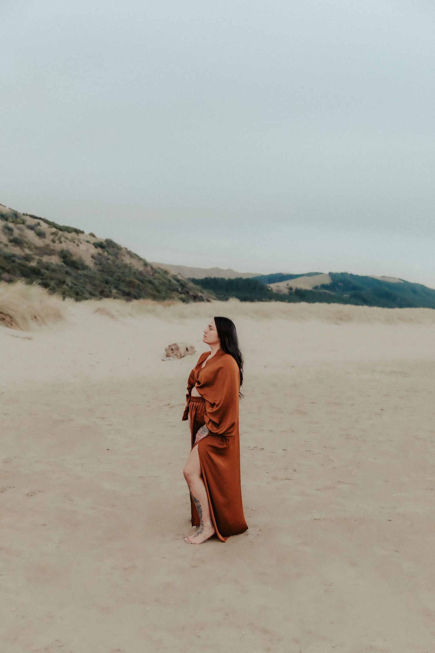 Figure in rust-colored dress poses gracefully on sandy beach with mountains in background.