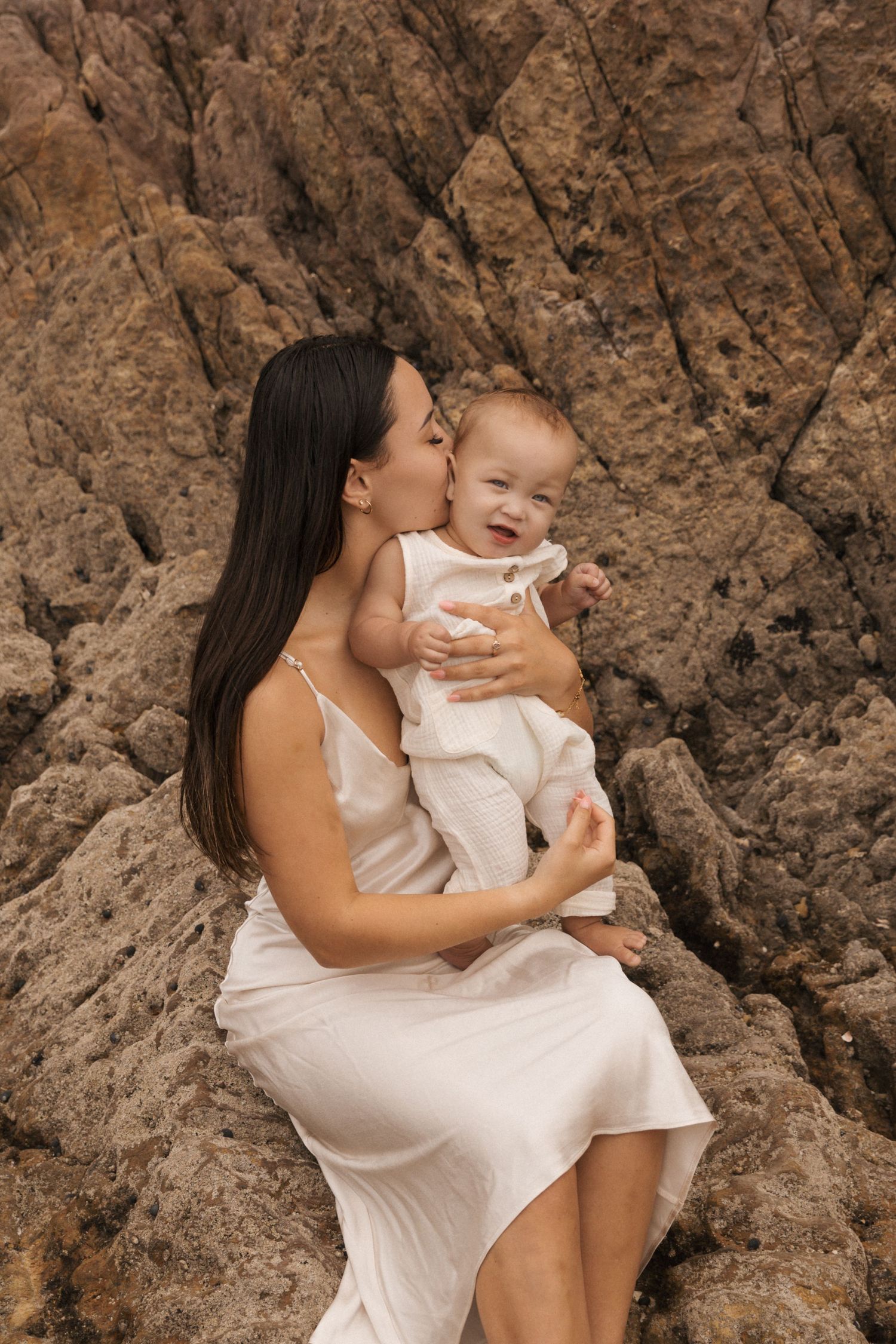 Mother and infant sharing tender moment against rocky beach backdrop.