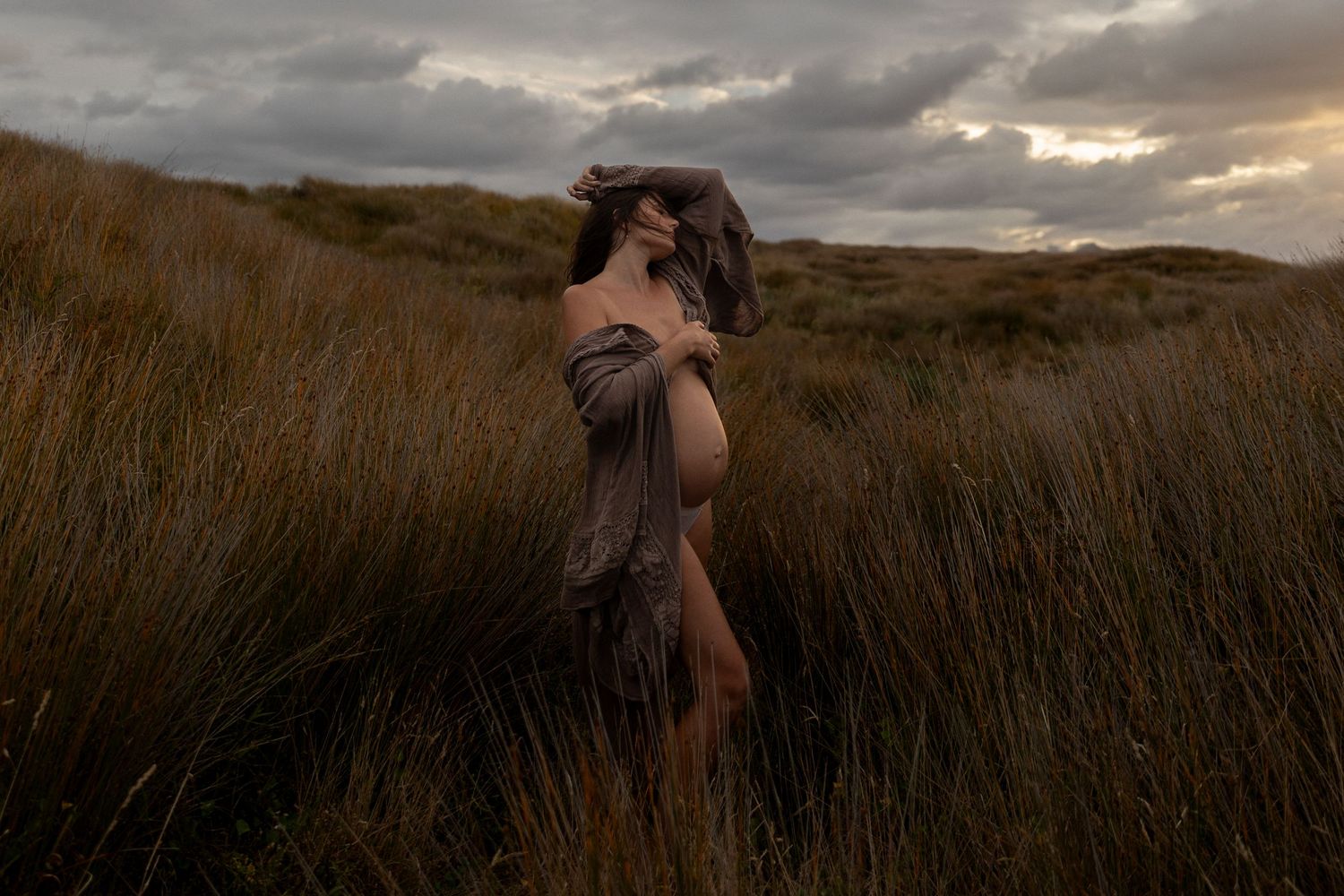 Dramatic maternity portrait against stormy sky with golden grass in foreground.