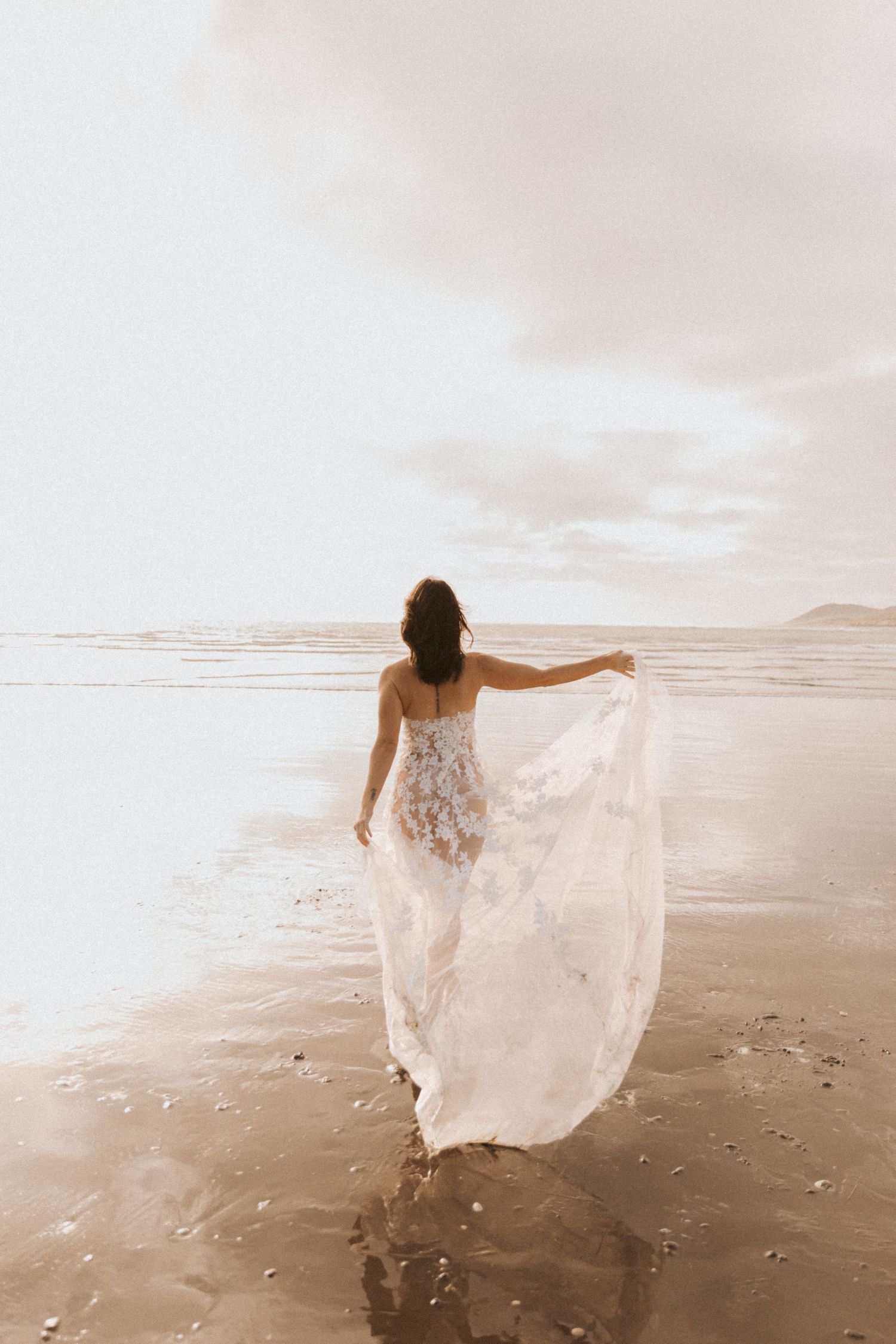 Woman twirls in sheer white lace dress on sandy beach at dusk.