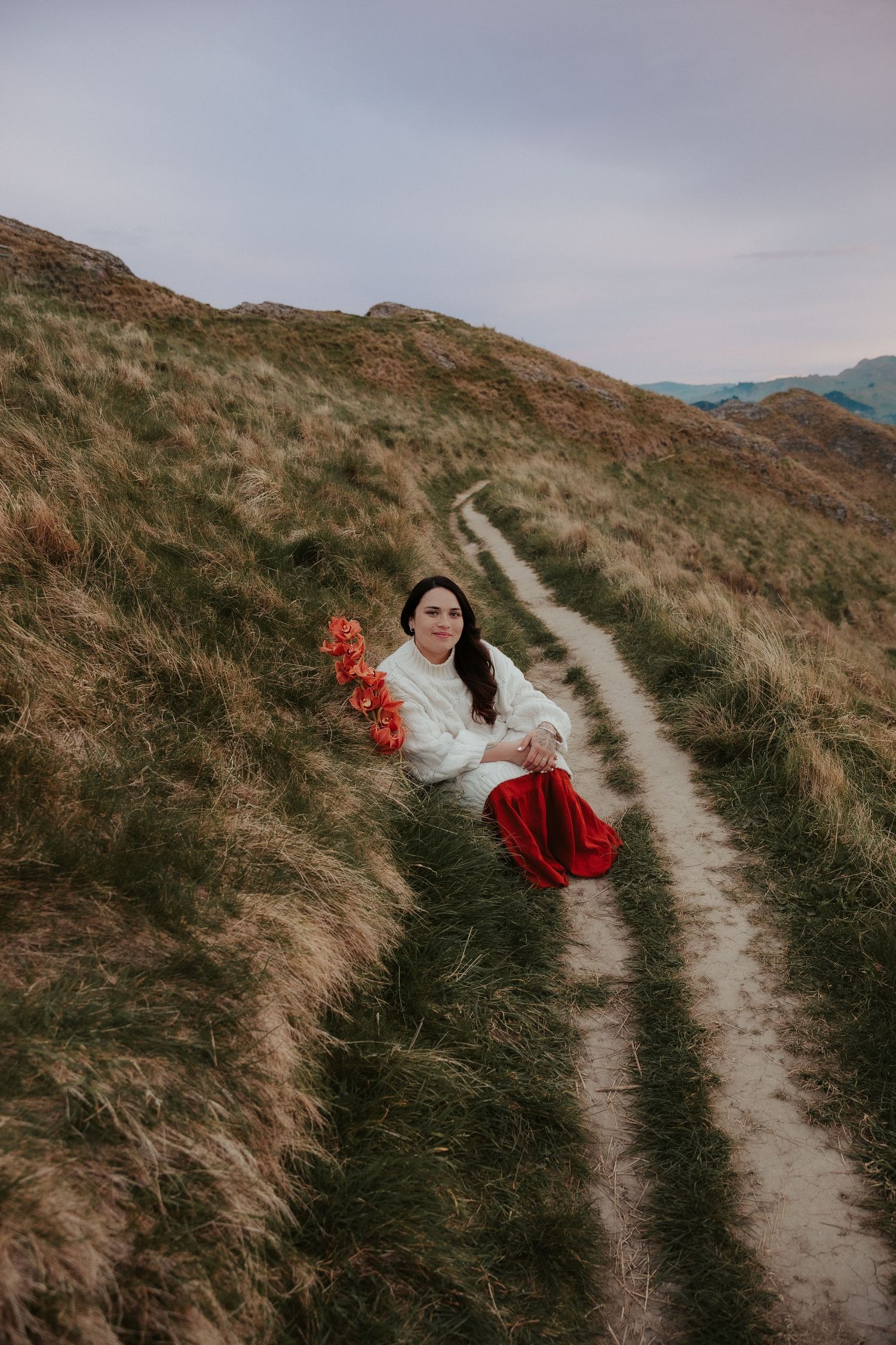 Person in white dress with red flowers on winding dirt path through coastal hills.