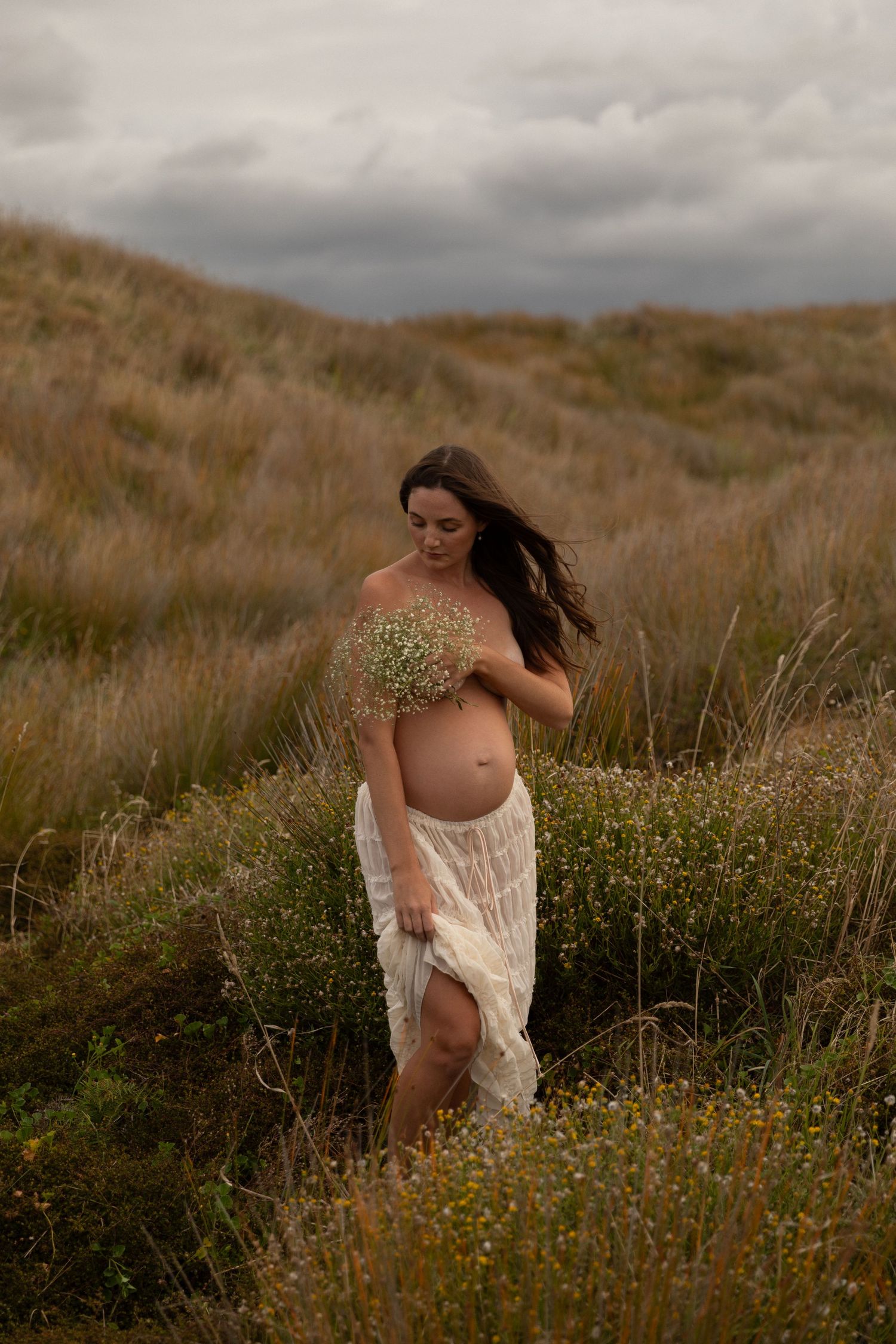 Expectant mother poses gracefully among wildflowers in flowing white fabric.