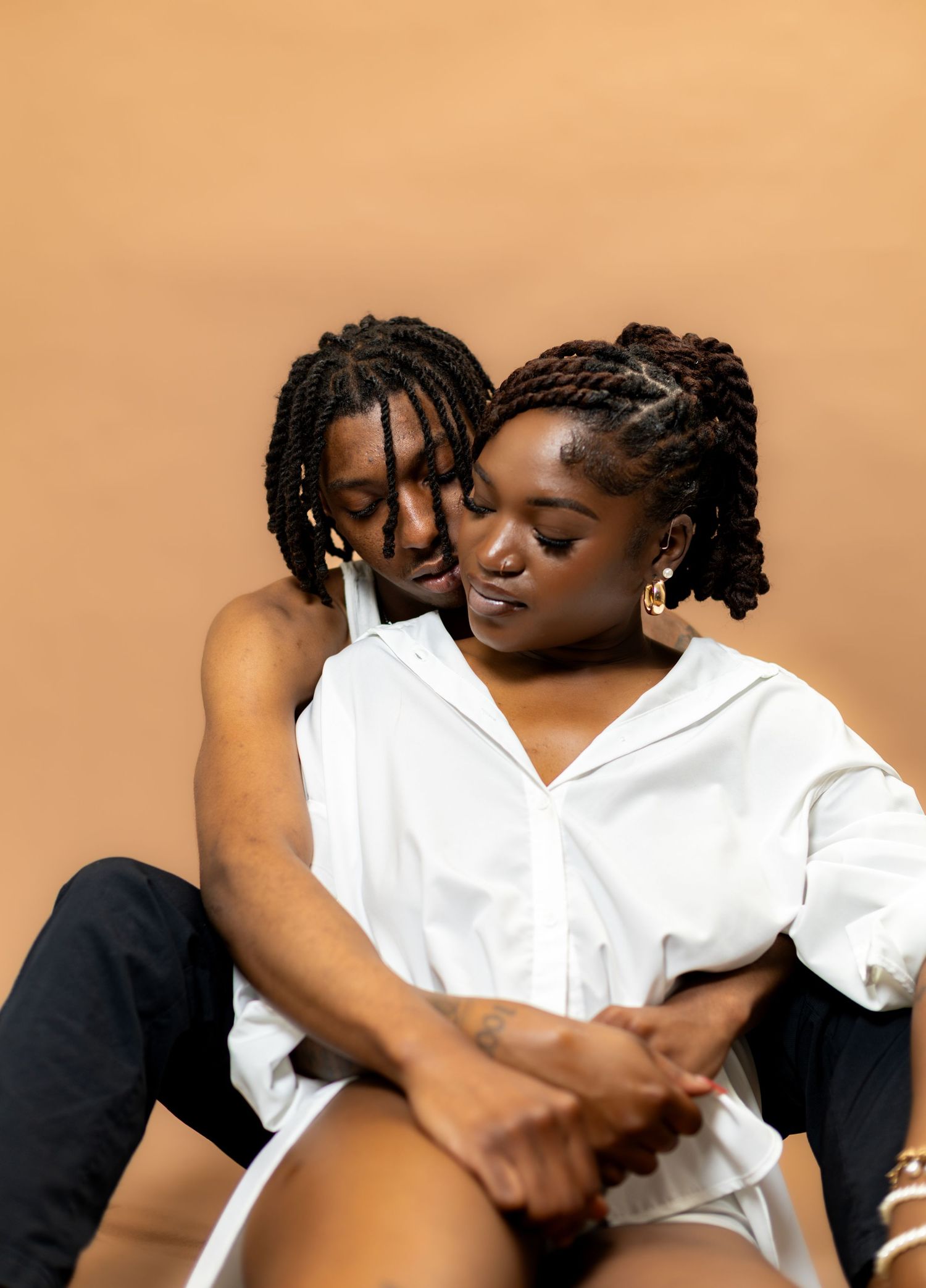 A couple shares an intimate embrace, sitting together against a warm beige background in coordinating white and black outfits.