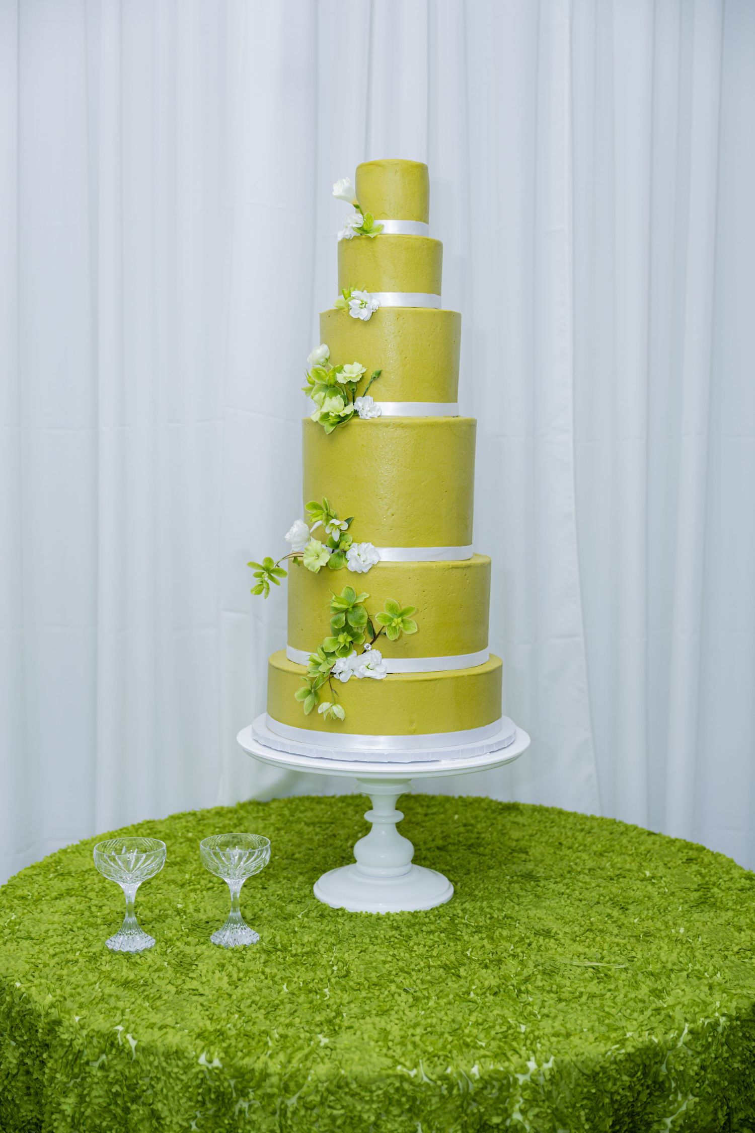 Elegant six-tier green wedding cake with white floral accents on a white stand, displayed on a green tablecloth.