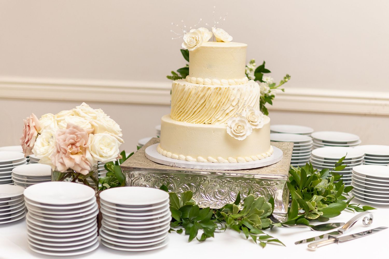 Elegant white wedding cake with stacked plates and greenery displayed on a dessert table at a reception.