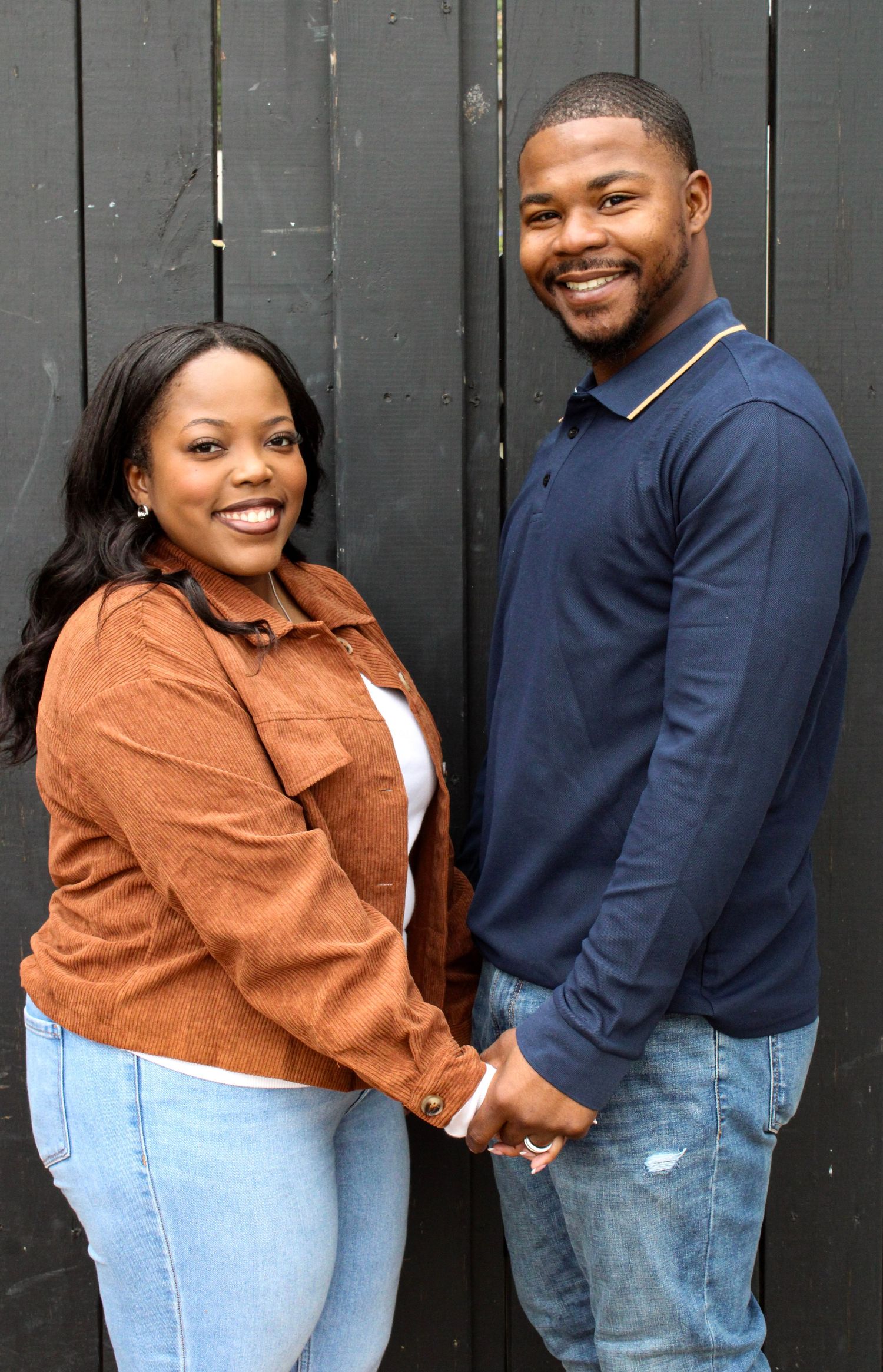 A couple poses against a dark fence, with one wearing a brown jacket and the other in navy blue.