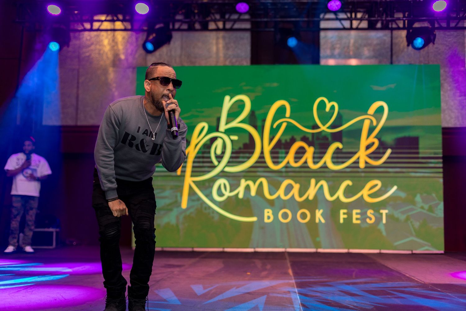 Man speaking on stage at Black Romance Book Fest event with colorful LED backdrop lighting.
