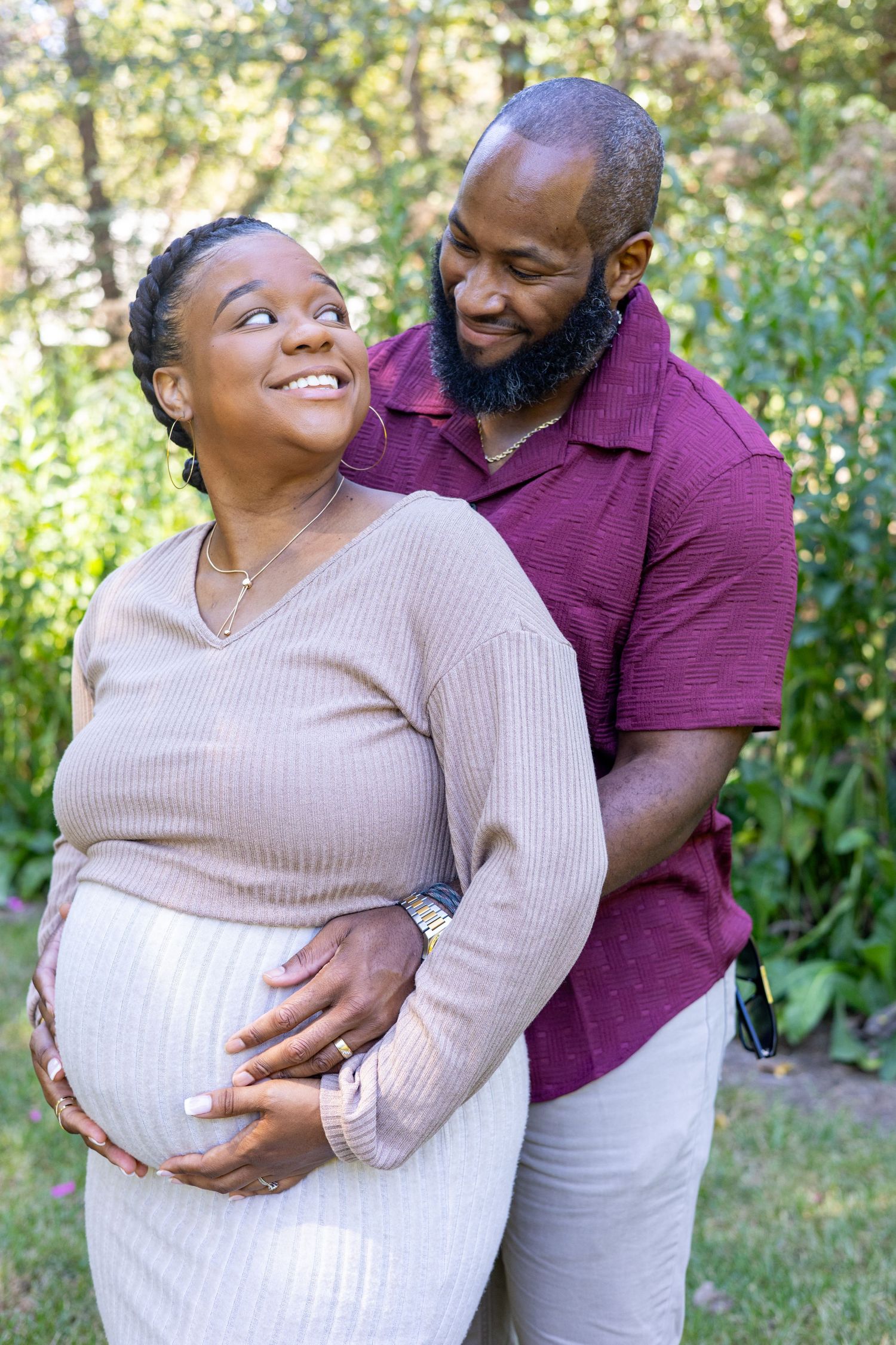 A couple embraces in a garden setting while wearing beige and burgundy outfits.