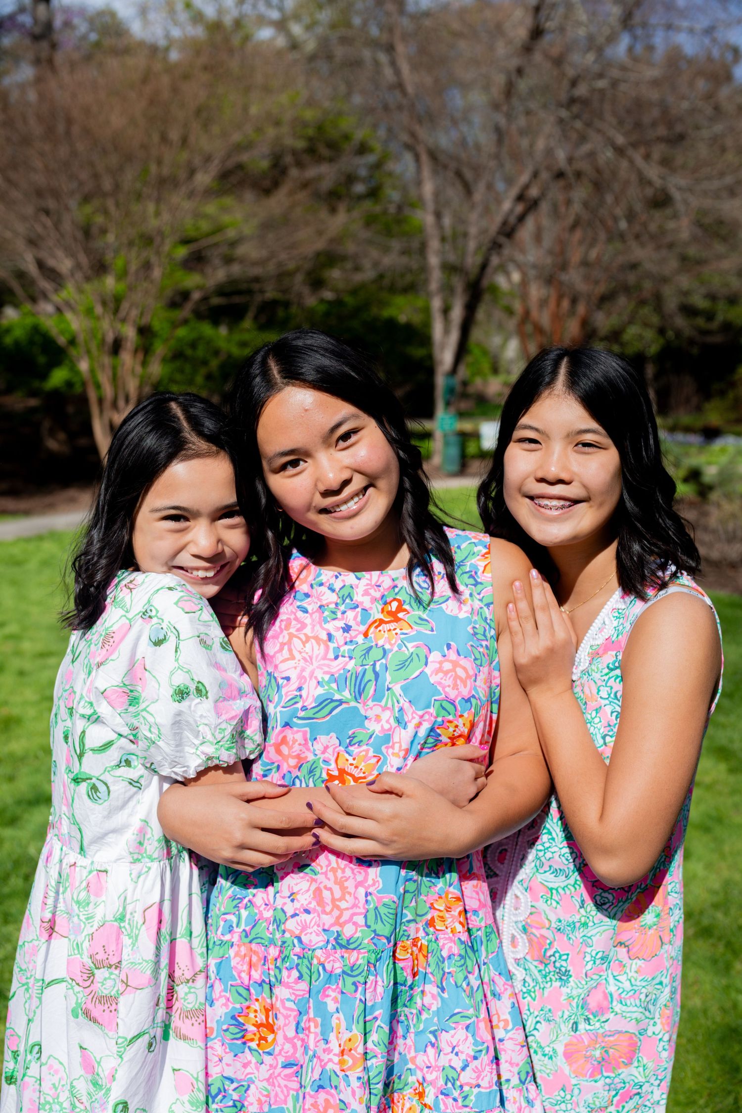 Three smiling girls in colorful floral dresses posing together outdoors in a sunny green park setting.