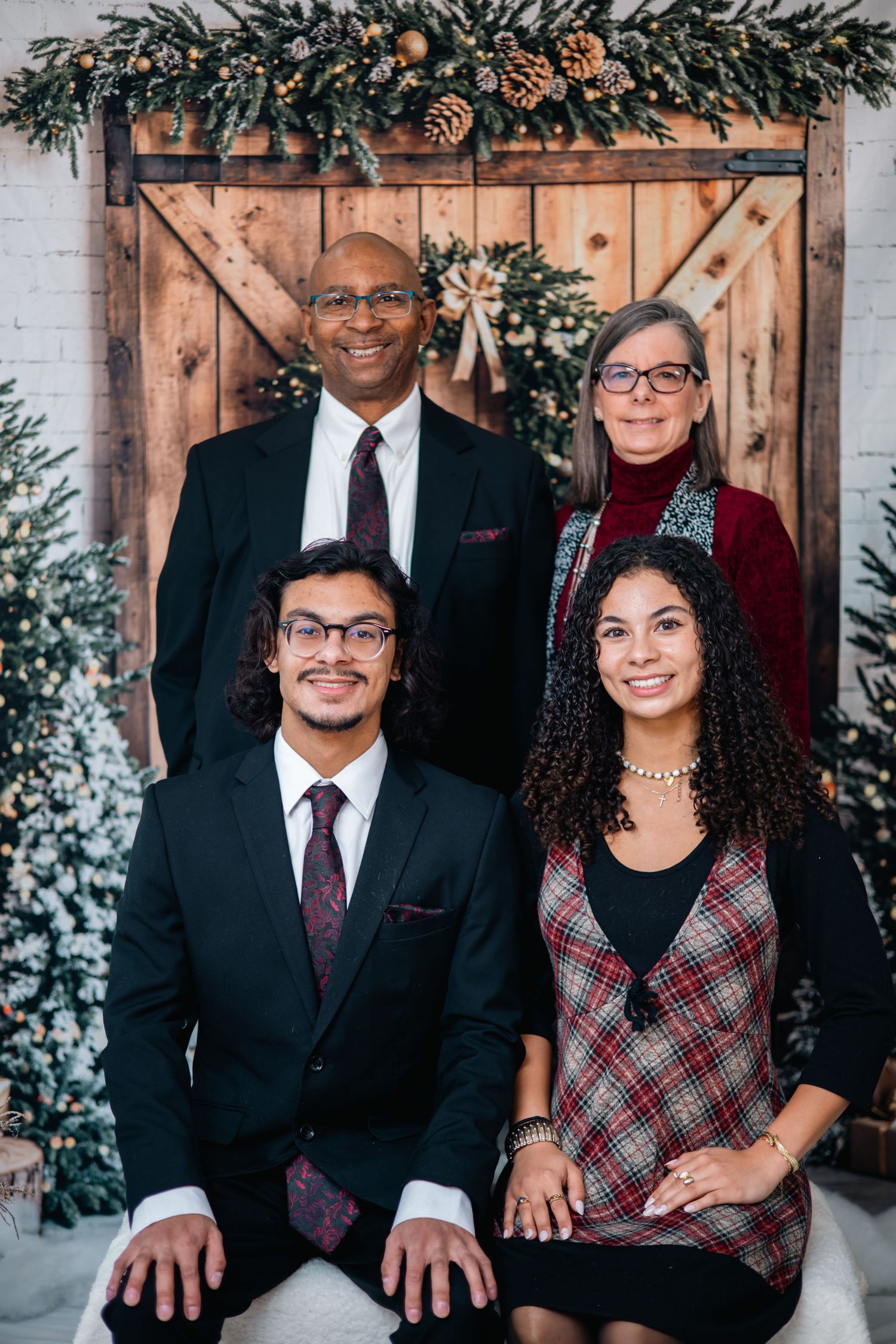 A family poses together for holiday portraits against a rustic wooden backdrop decorated with pine garland and pinecones.