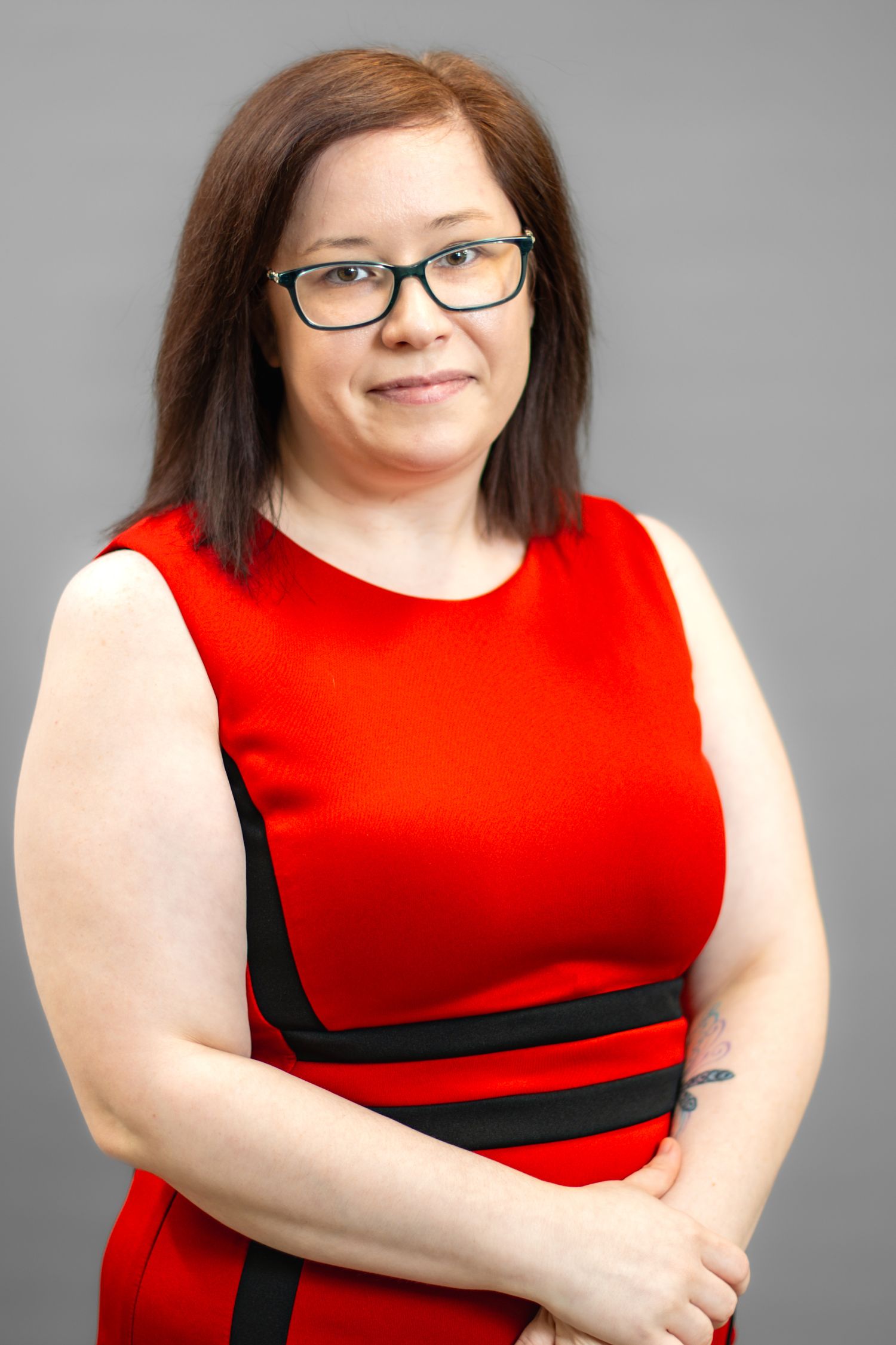 Woman in red sleeveless dress with black trim, wearing glasses, brown hair, arms crossed, gray background.