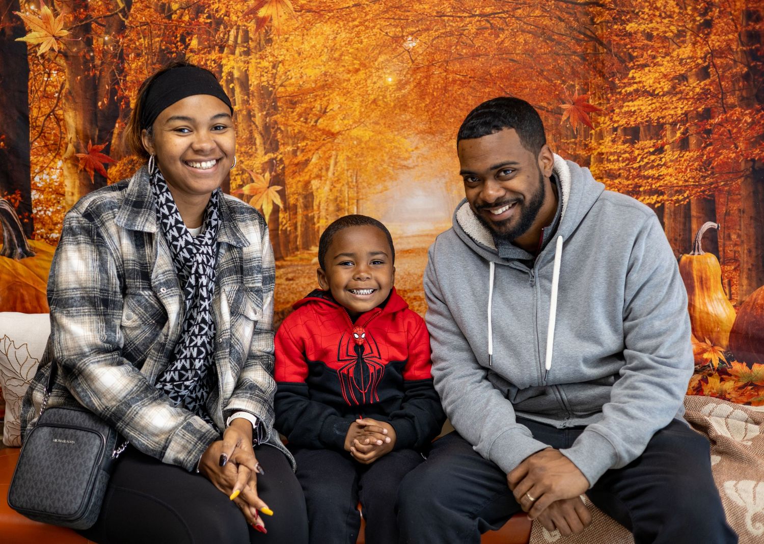 A happy family sits together outdoors during autumn with vibrant orange fall foliage in the background.