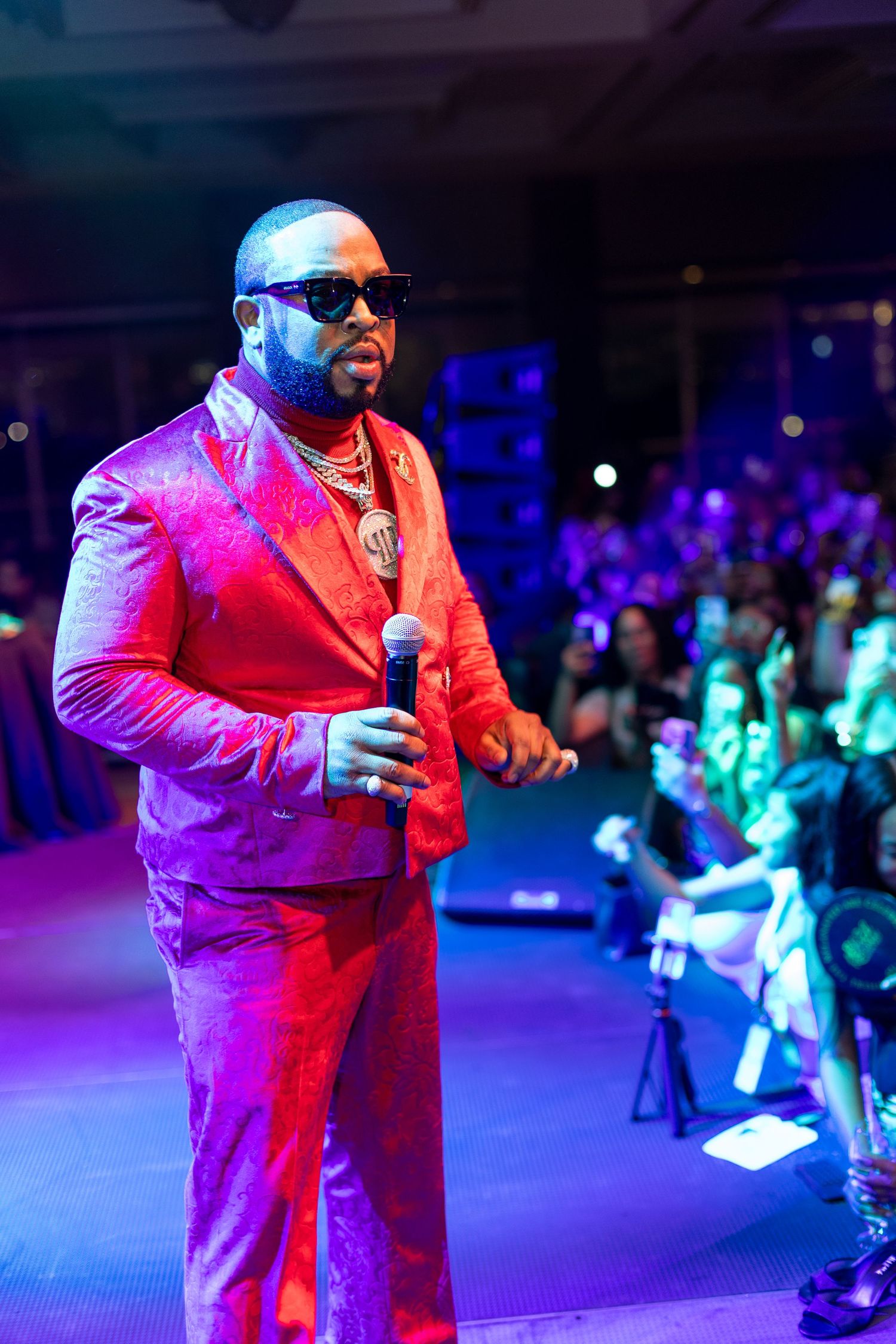 Man in red suit and sunglasses holds microphone on stage under colorful lights at a live event.