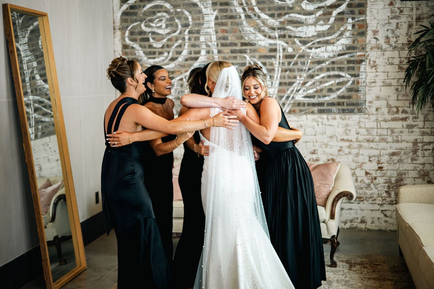 Wedding party in black dresses helps bride get ready in rustic brick room with illuminated wall decorations.