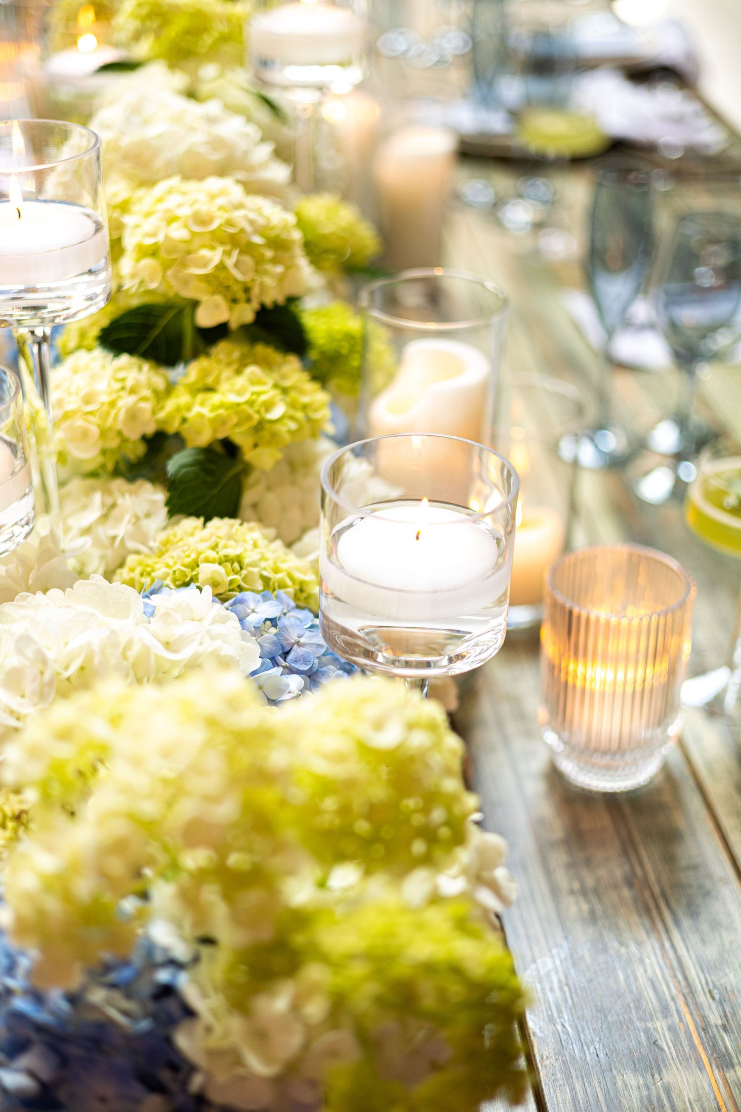 Wedding table decor with hydrangeas and glowing candles in glass holders on wooden surface.