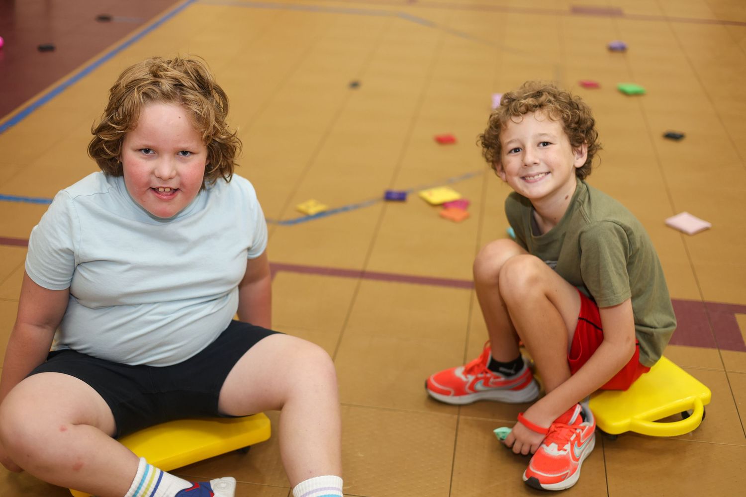 Two kids sitting on yellow scooter boards in a gym with scattered items on the floor.
