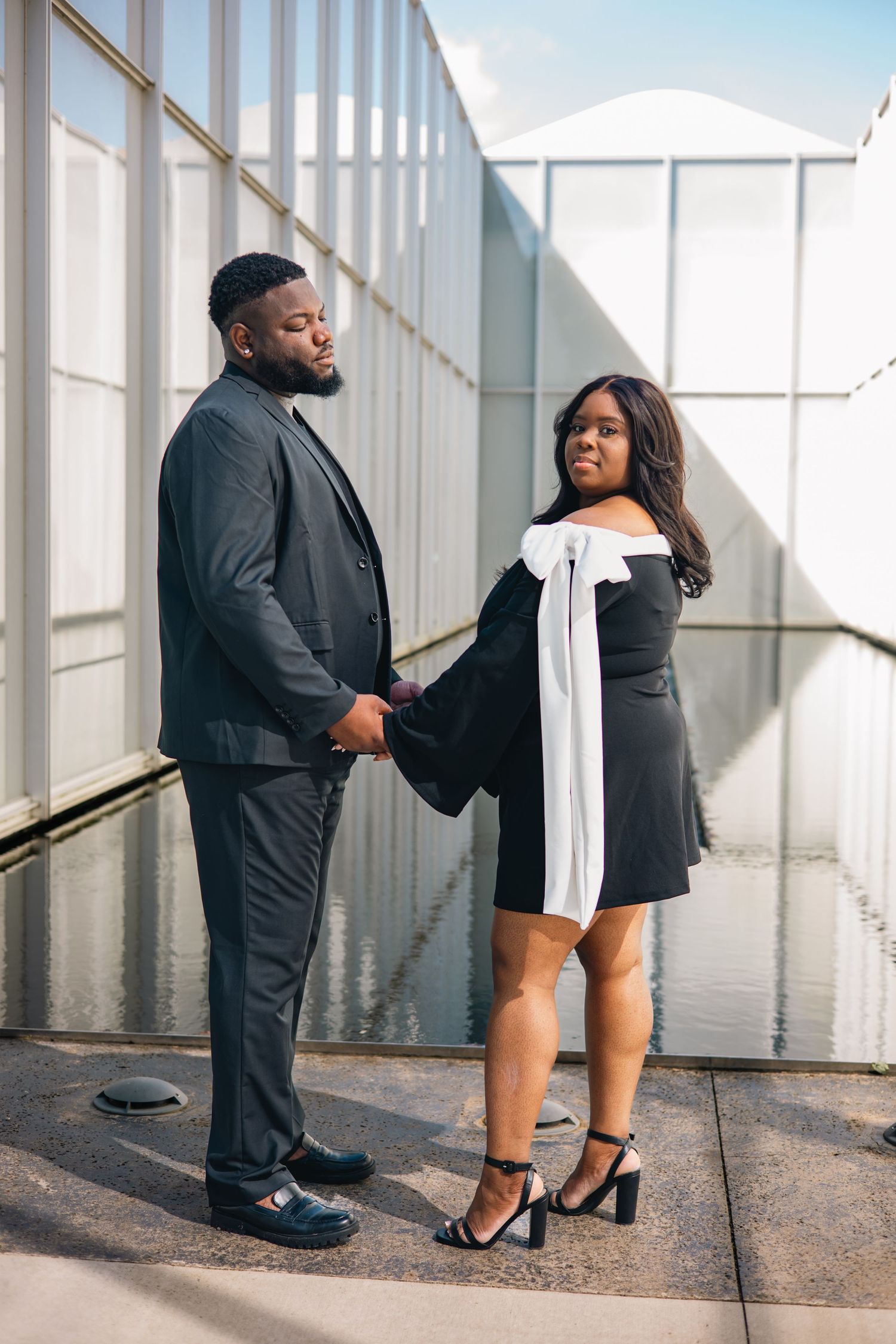 A couple in formal attire poses together in a bright hallway with white walls and natural lighting.