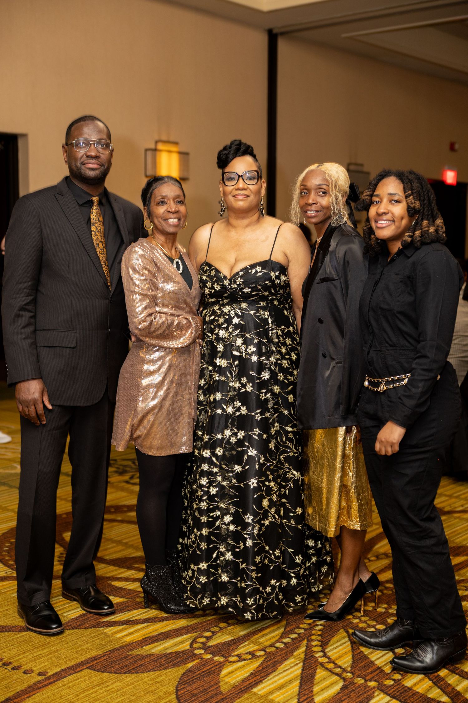 A group of people in formal attire pose together at an indoor event with golden carpeted floors.