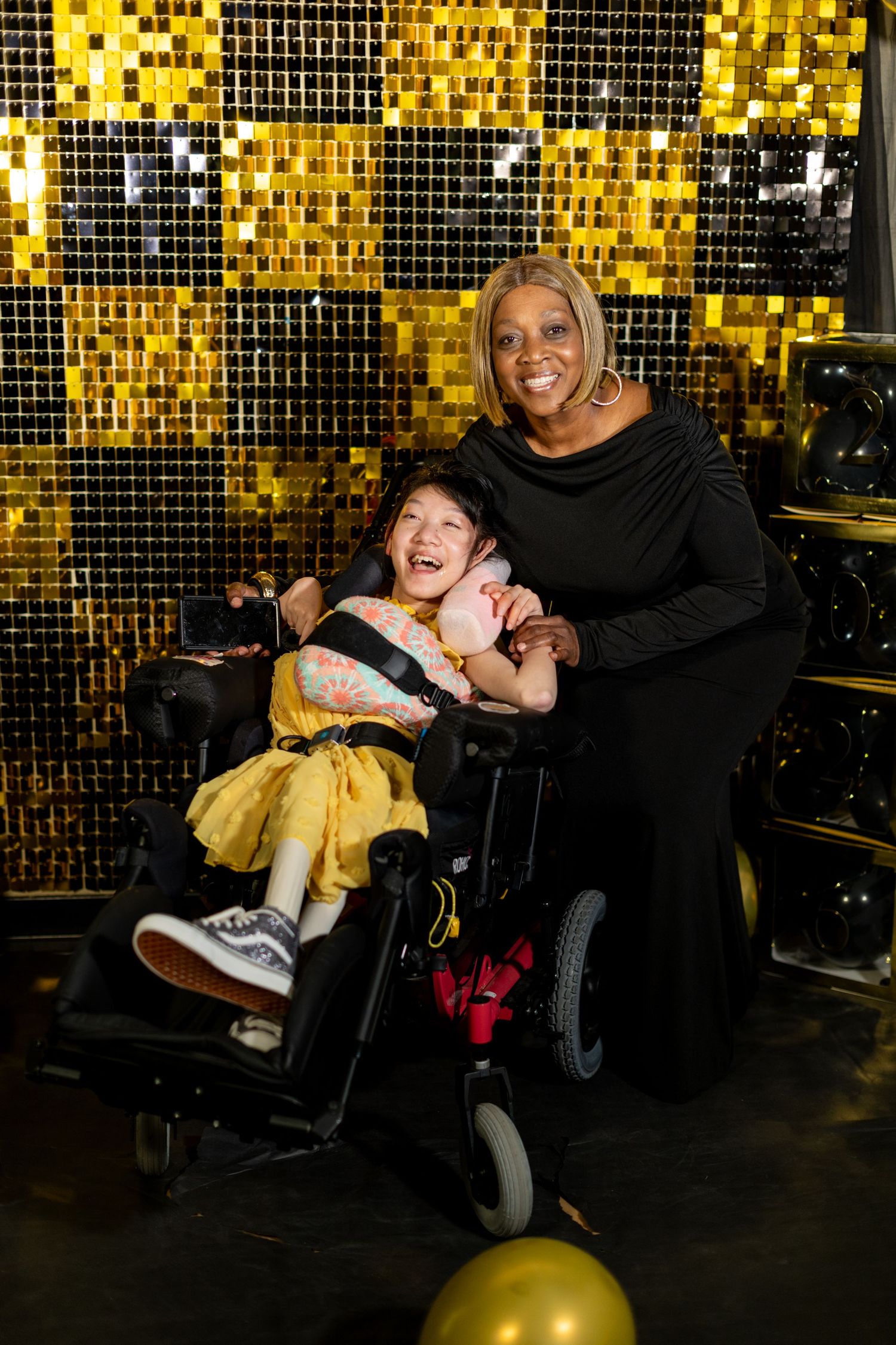 A smiling child in a wheelchair wearing yellow poses with a woman in black against a gold sequin backdrop.