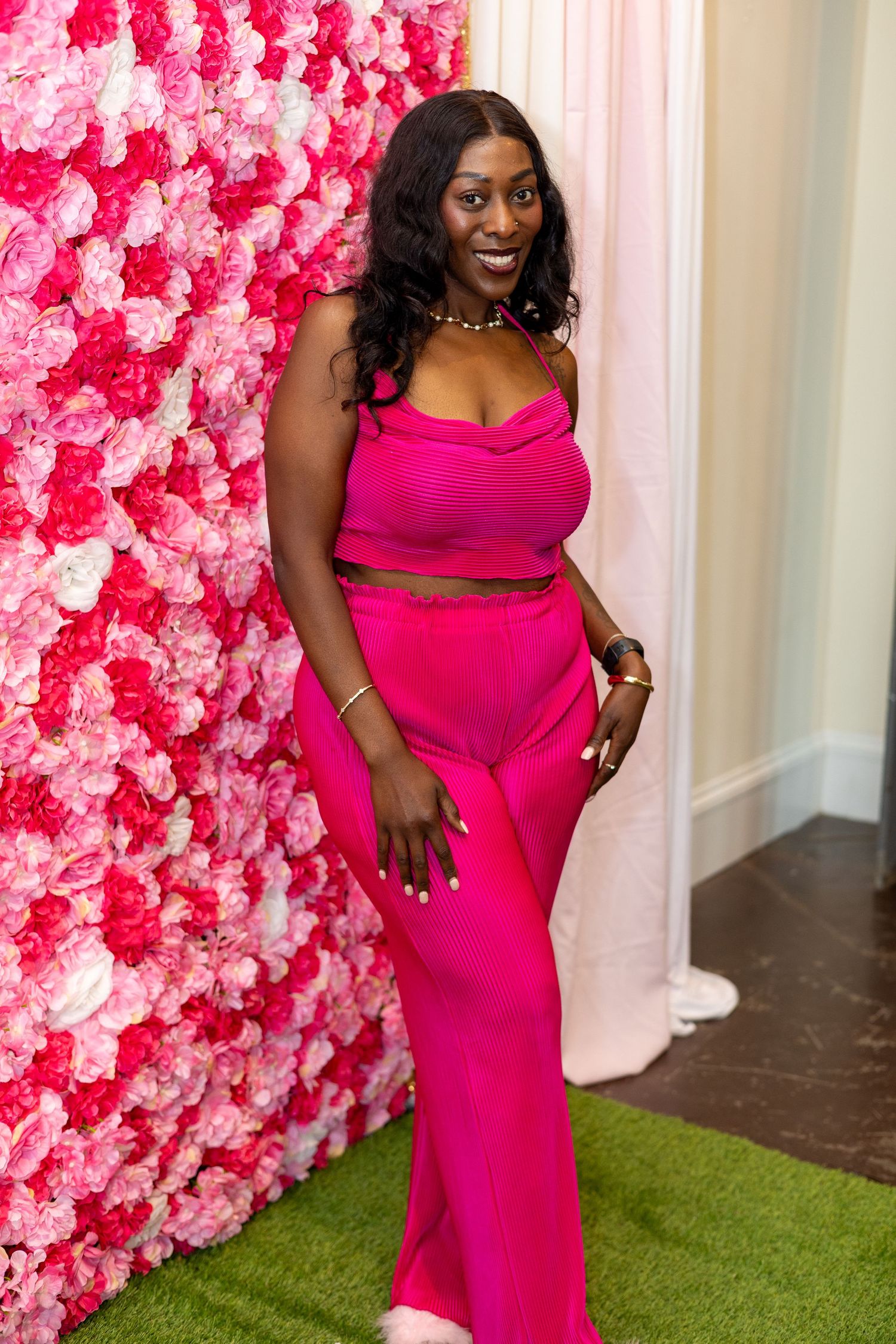 A person in a vibrant hot pink dress poses against a pink floral wall backdrop on artificial grass.
