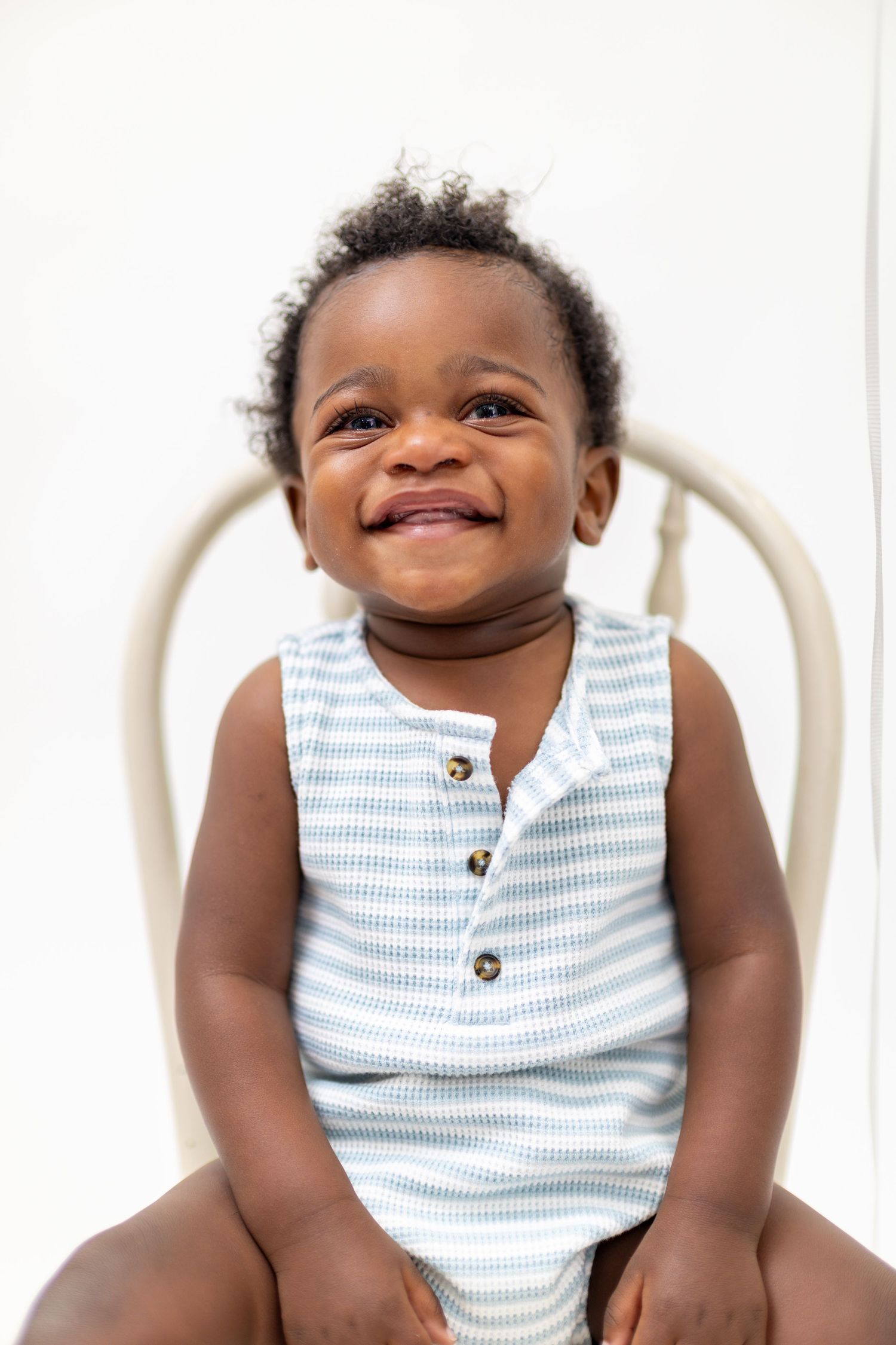 Smiling Black baby boy sitting on a chair wearing a blue striped sleeveless romper with buttons.