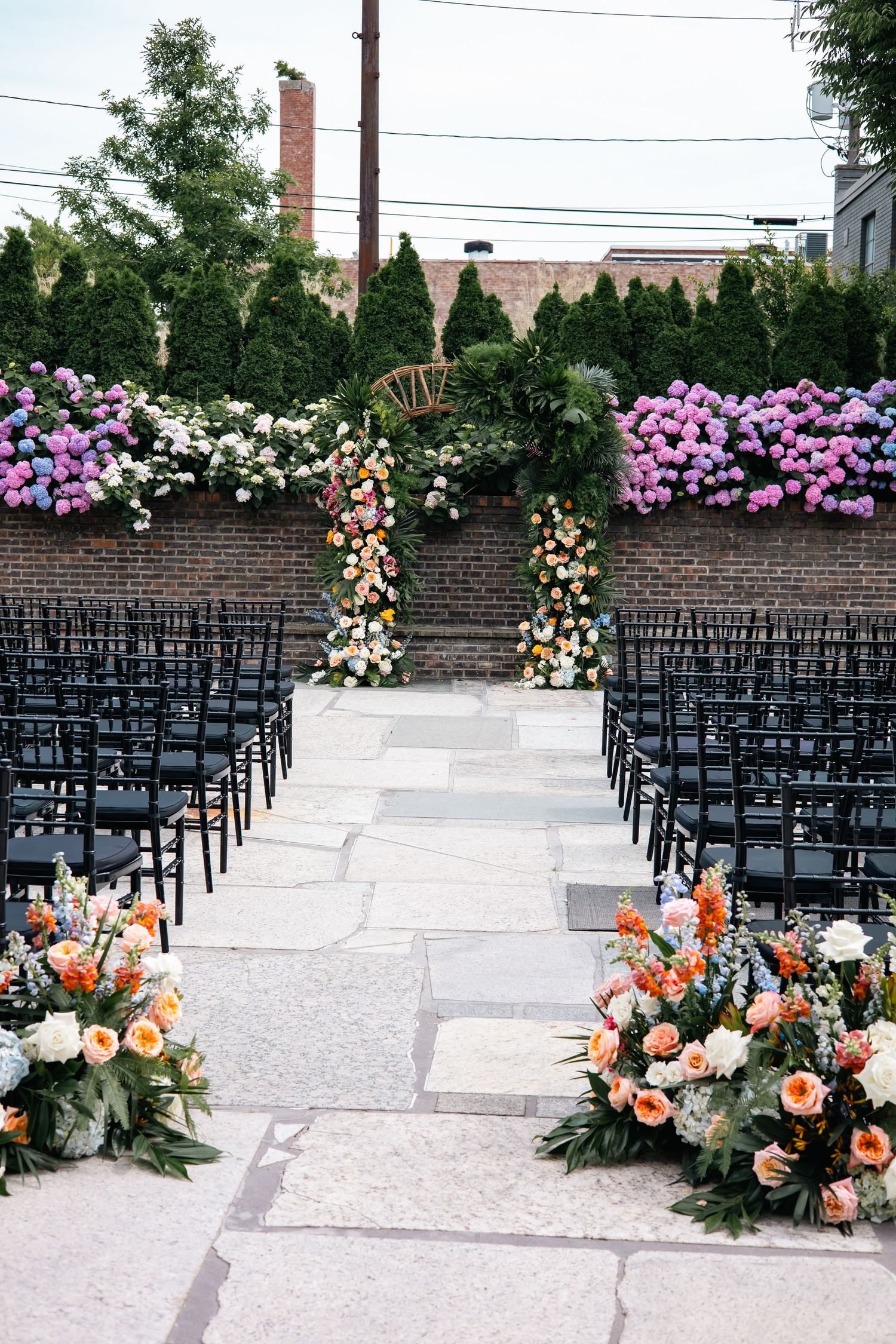 Outdoor wedding ceremony setup with rows of black chairs and colorful floral arrangements along the aisle.