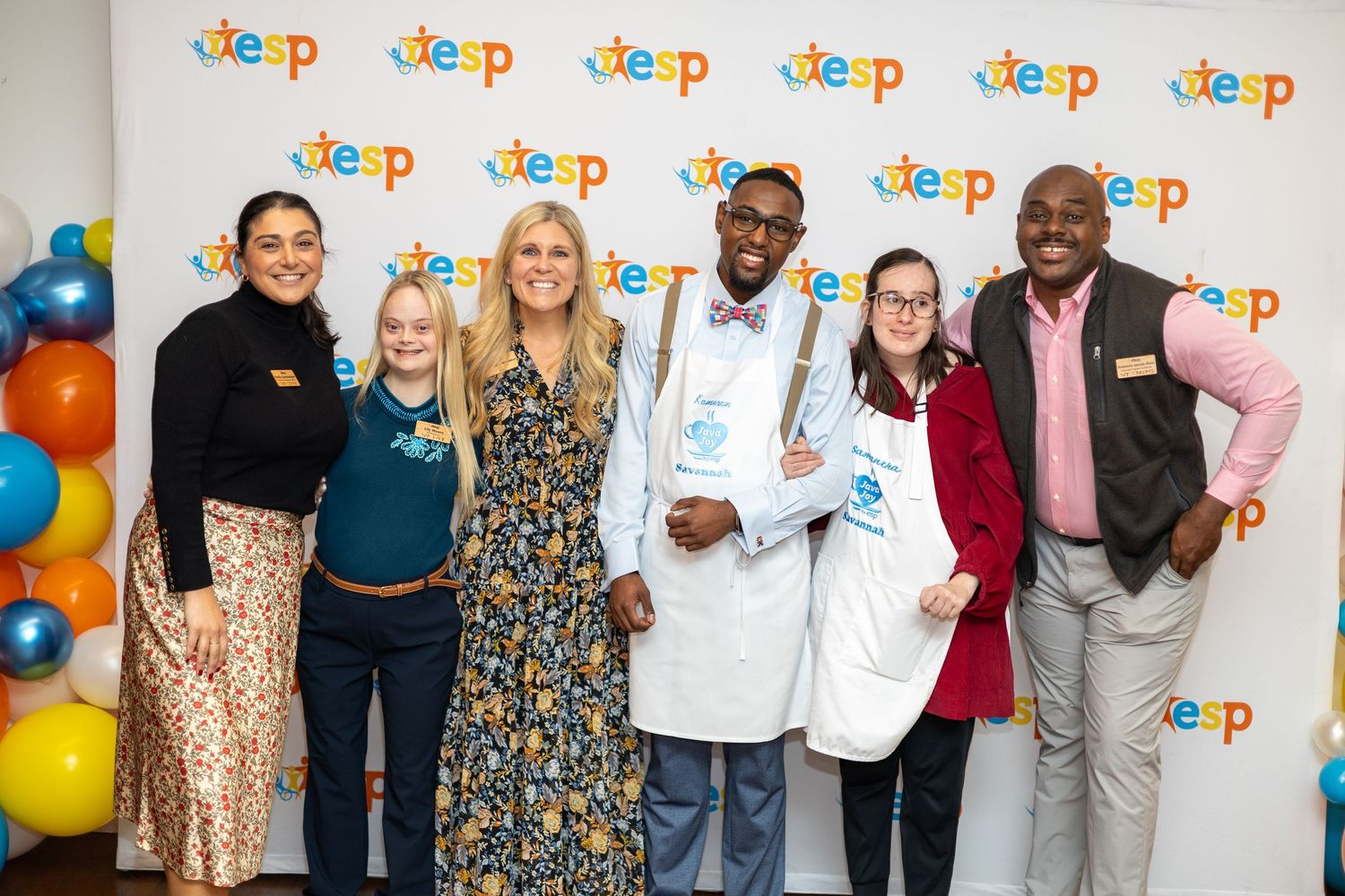 Group of six people posing together at a TESP event, smiling in front of a branded backdrop with colorful balloons.