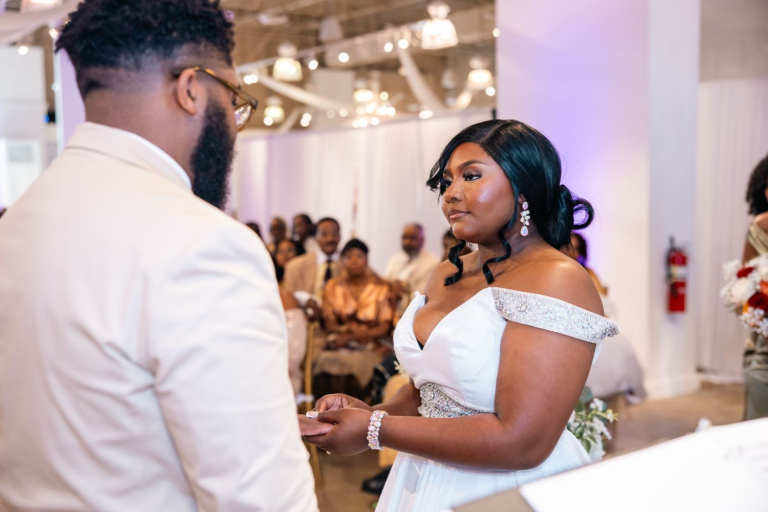 Bride in off-shoulder white gown exchanges vows with groom during elegant indoor wedding ceremony.