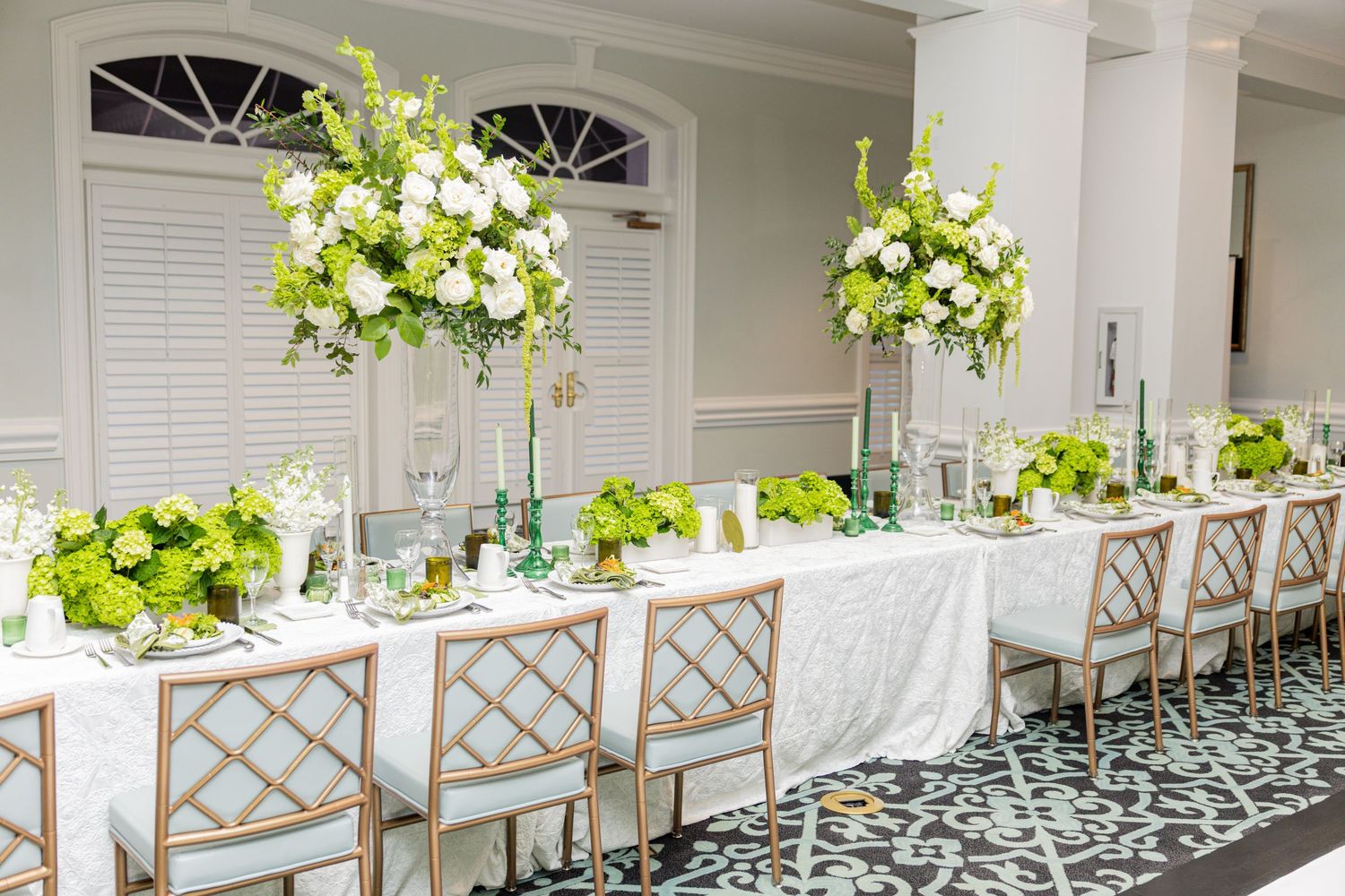 Elegant wedding reception table with tall green and white floral centerpieces, white linen, and gold bamboo chairs.