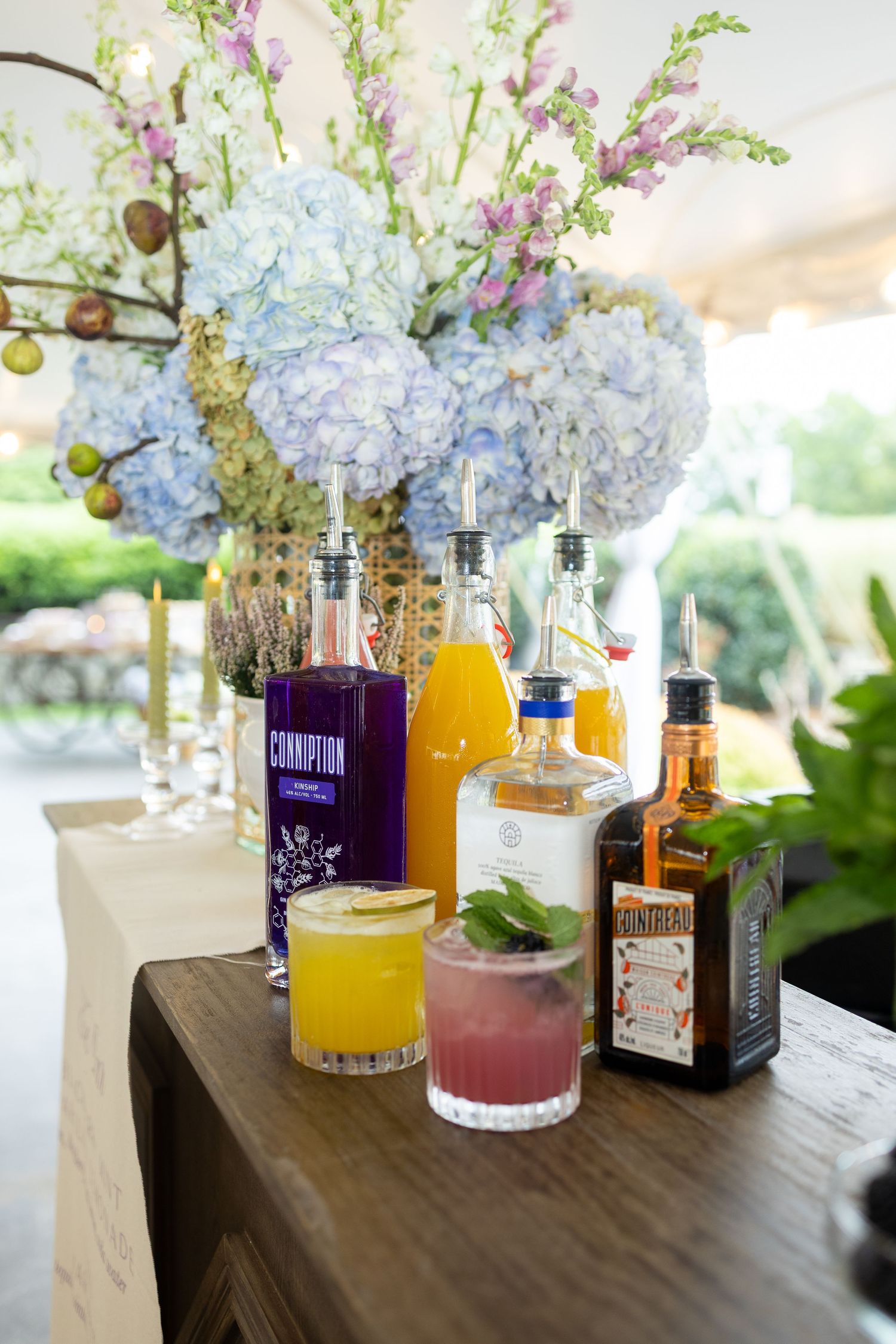 Assorted liquor bottles and colorful cocktail mixers arranged on a wooden table with blue floral decorations.