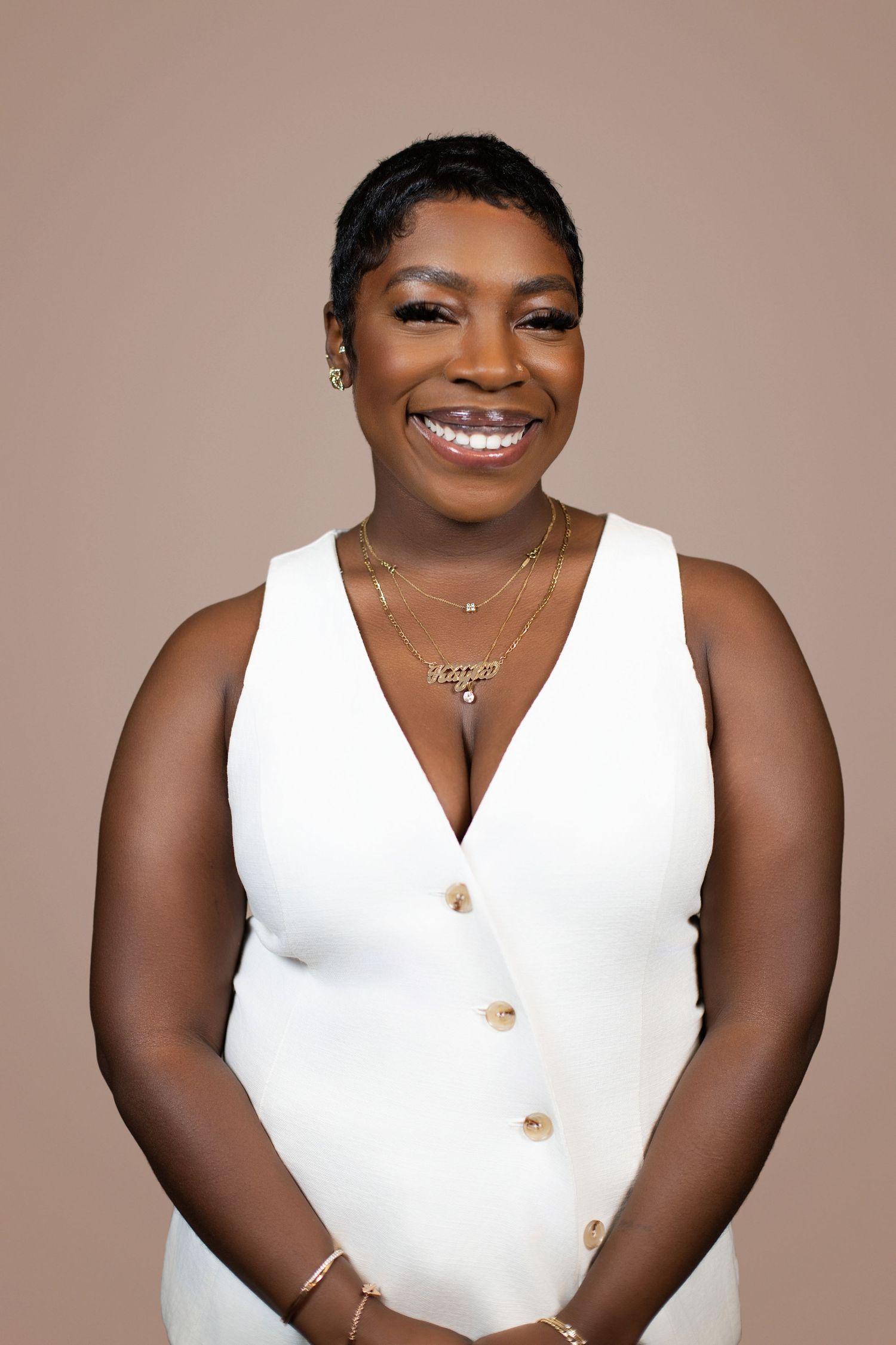 Smiling woman in white sleeveless dress with gold jewelry poses confidently against a warm beige background.