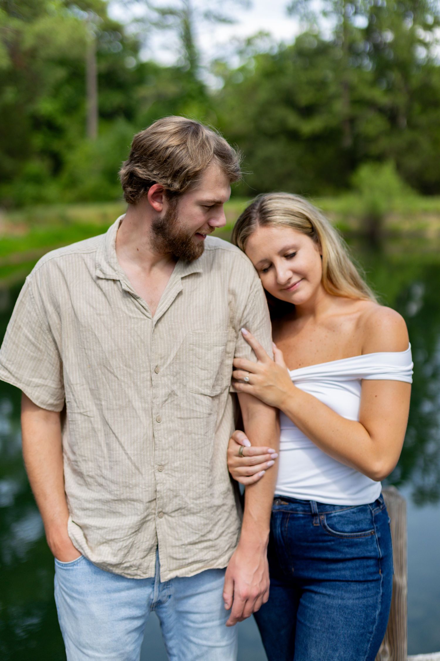 A couple stands together near a serene pond, the woman in a white off-shoulder top leaning on a bearded man in a beige shirt.