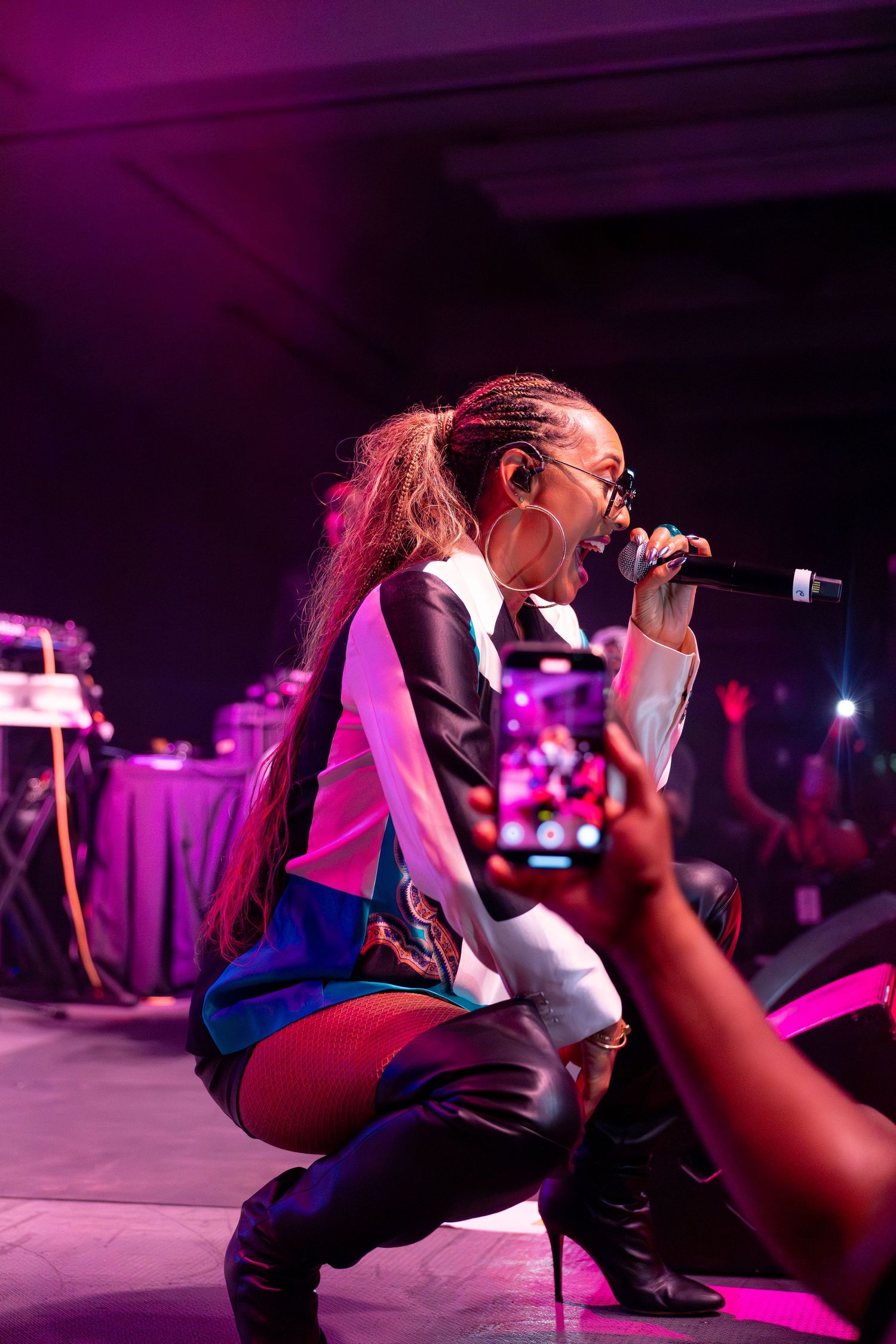 Female performer crouching on stage singing into microphone under purple lights while fan records with smartphone.