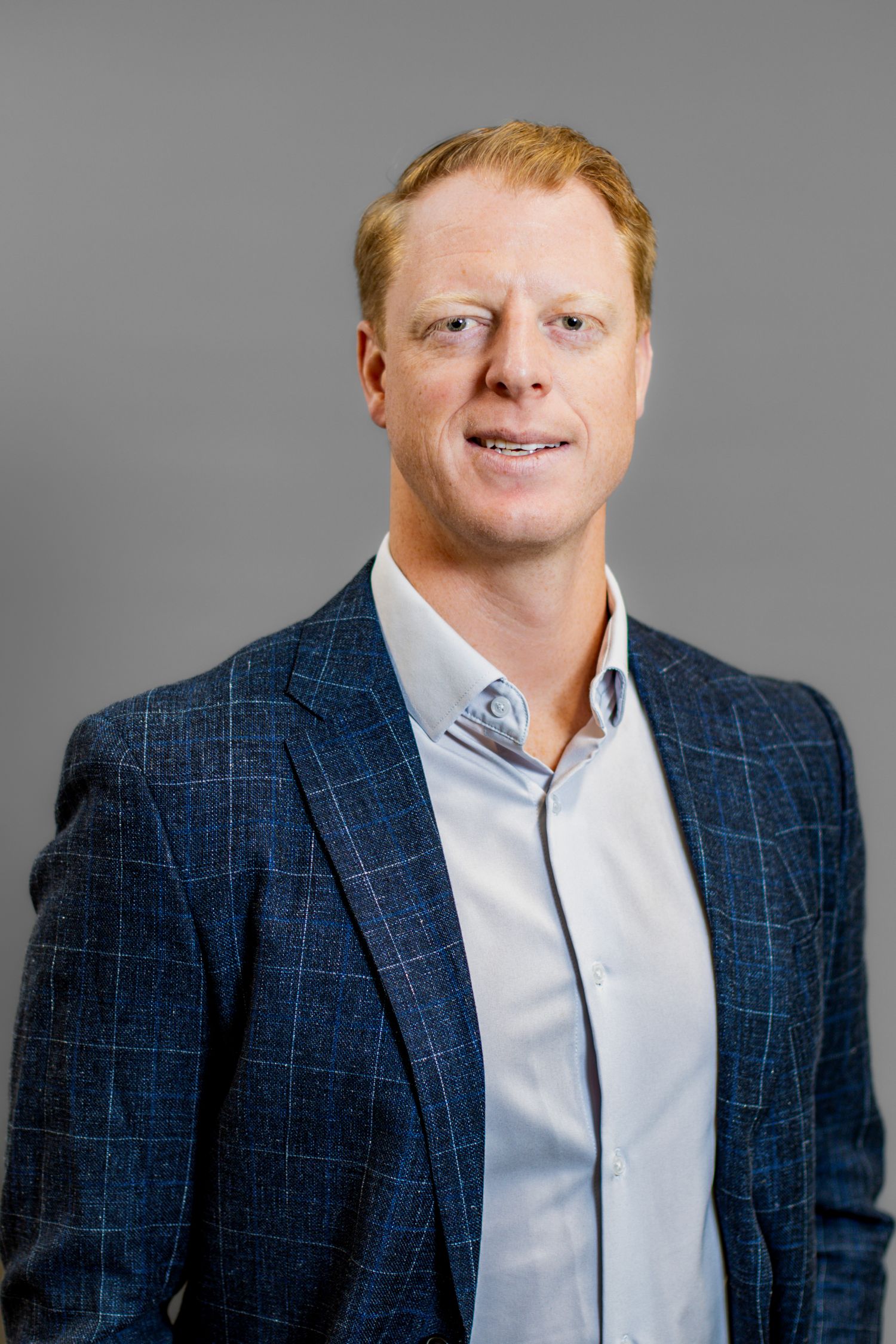 Professional headshot of a smiling red-haired man in a navy plaid blazer and light blue shirt against a gray background.