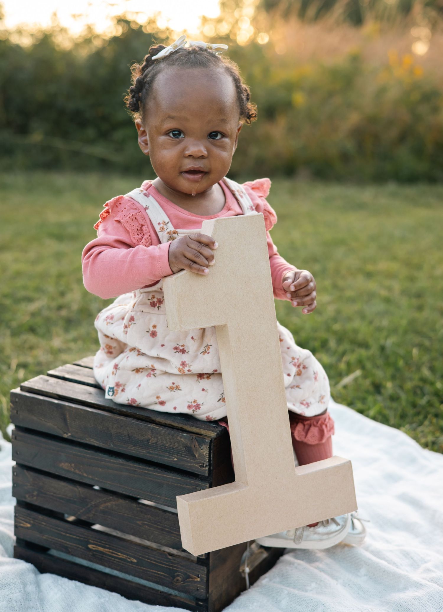 A small child in a pink outfit sits on wooden crates outdoors during golden hour, holding a decorative number one.