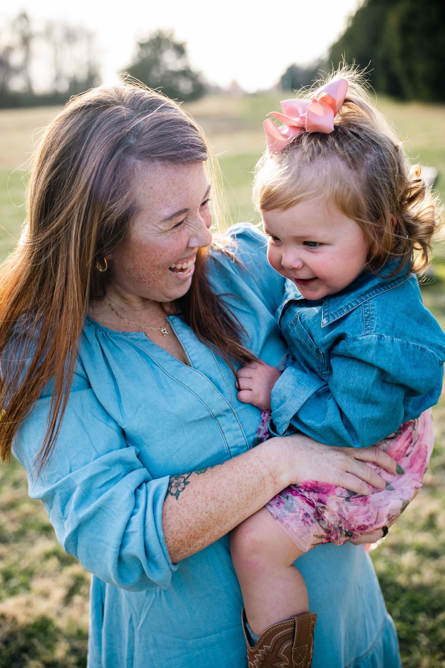 A joyful moment captured outdoors as a woman in blue holds a laughing toddler wearing a pink bow and cowboy boots.