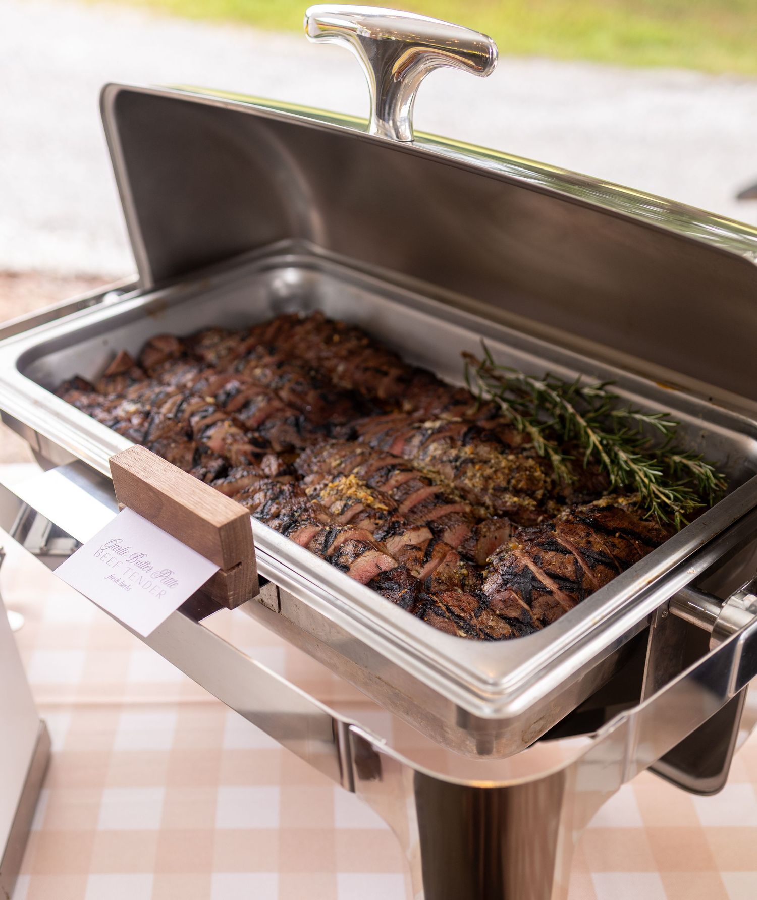 Row of stainless steel catering chafing dishes filled with grilled meats at a buffet service.