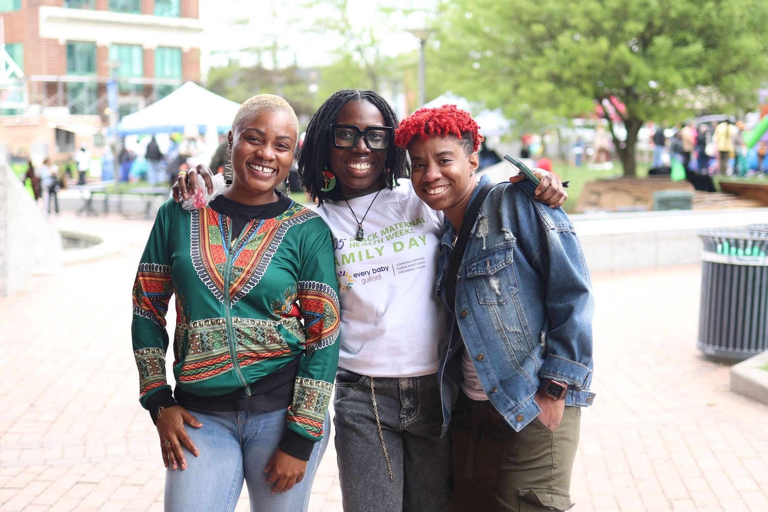 Three friends pose together smiling on brick walkway with buildings in background.