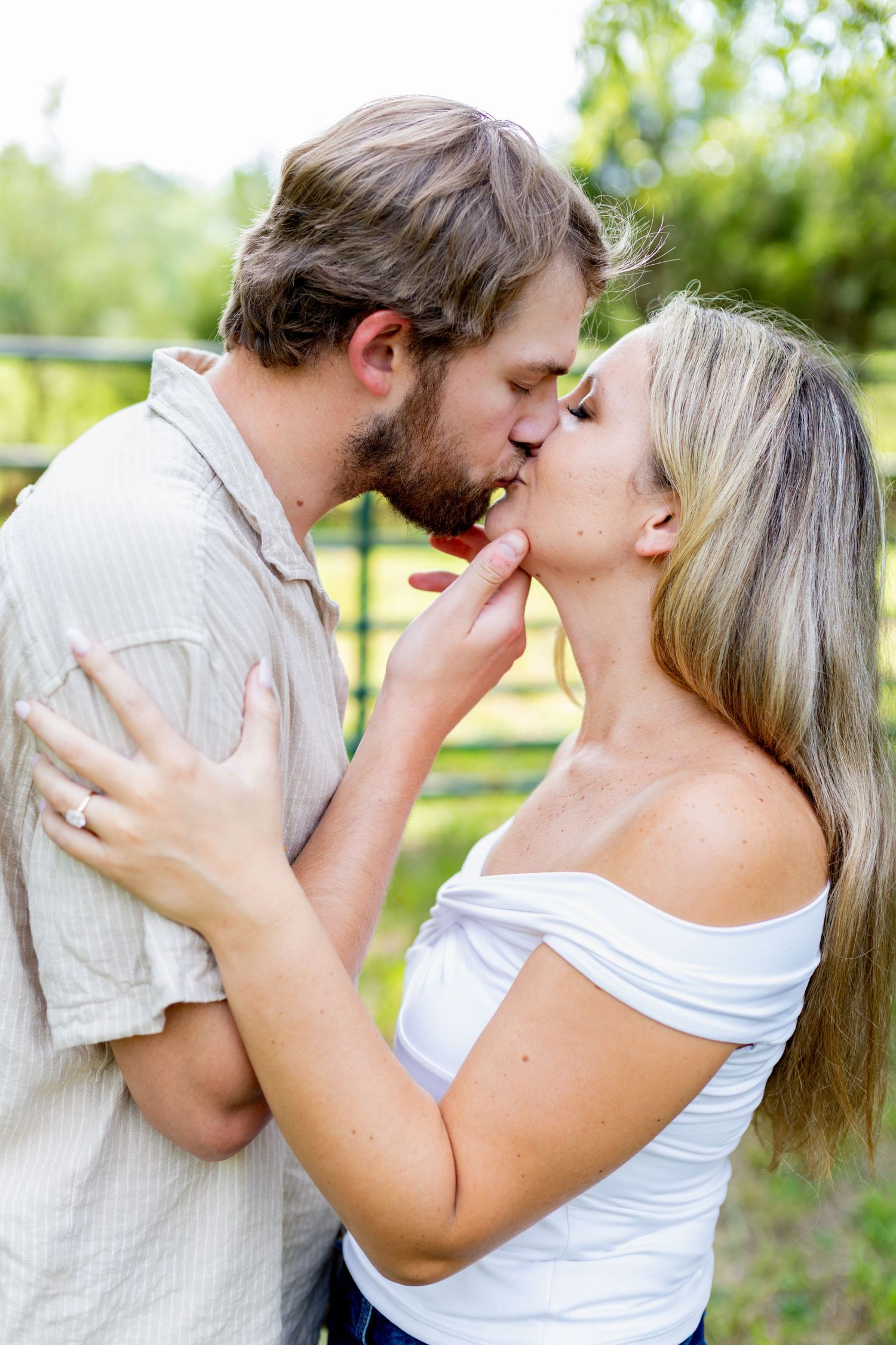 A couple shares a romantic kiss outdoors, with the woman wearing a white off-shoulder top and an engagement ring.