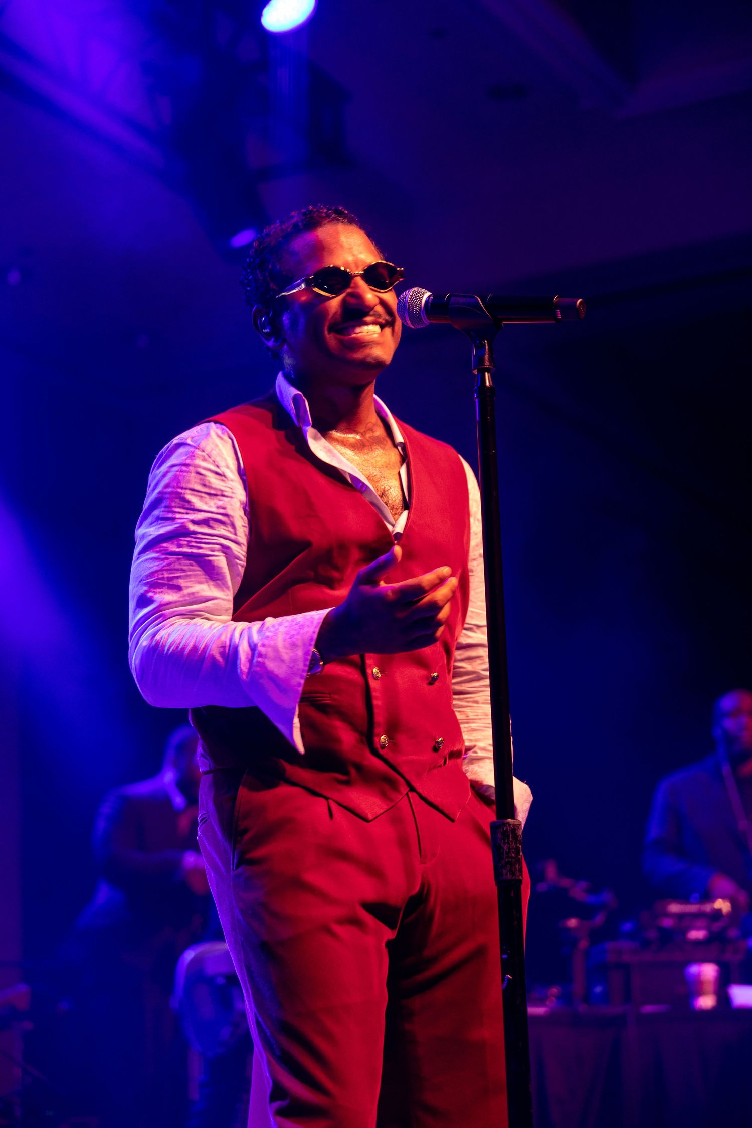 A smiling male performer in a red vest and sunglasses sings at a microphone on a purple-lit stage.