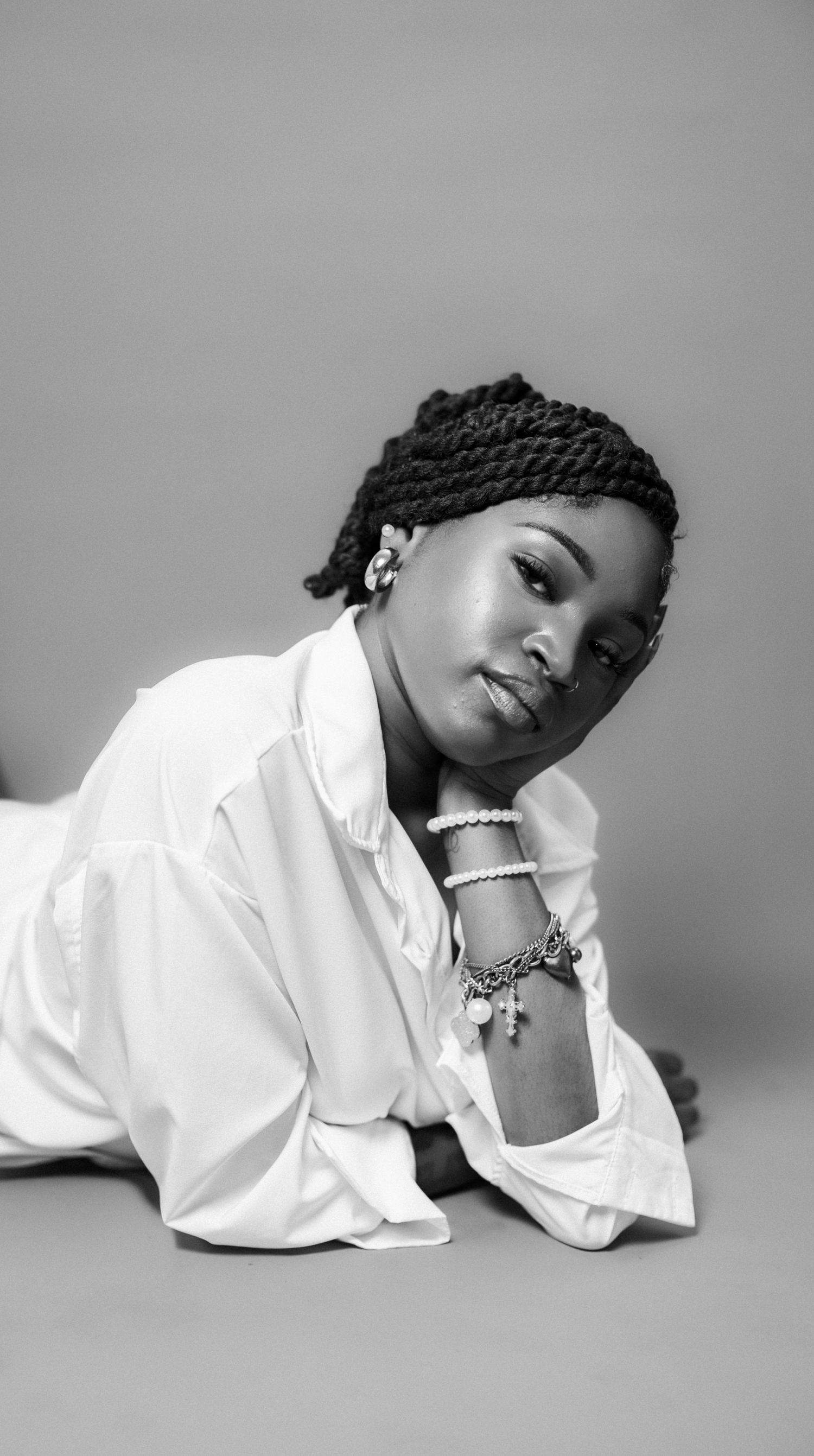 Black and white portrait of a young woman in a white shirt, braided hair, and charm bracelets, posing gracefully on the floor.