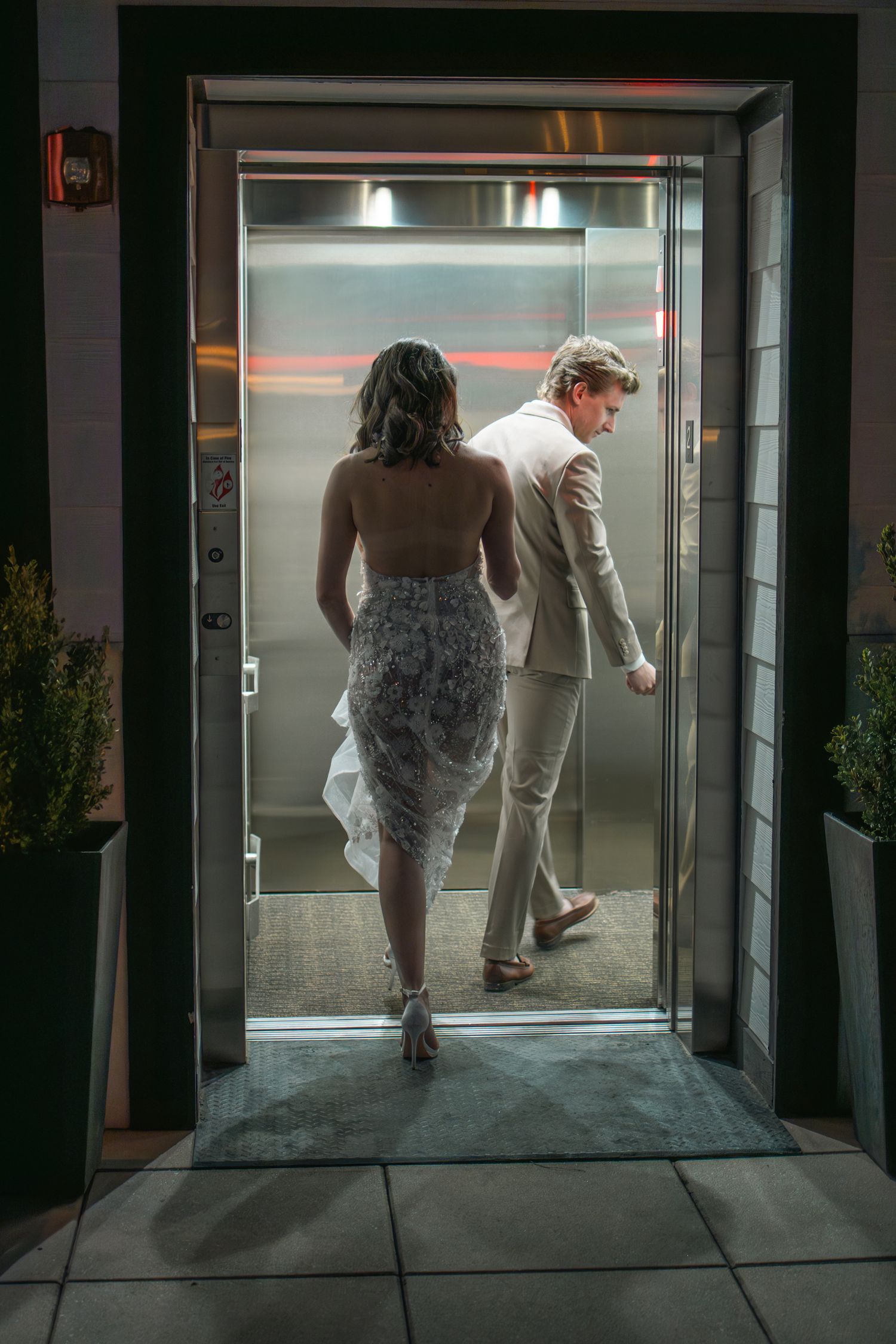 Two people in formal attire walk through an illuminated doorway in an evening setting.