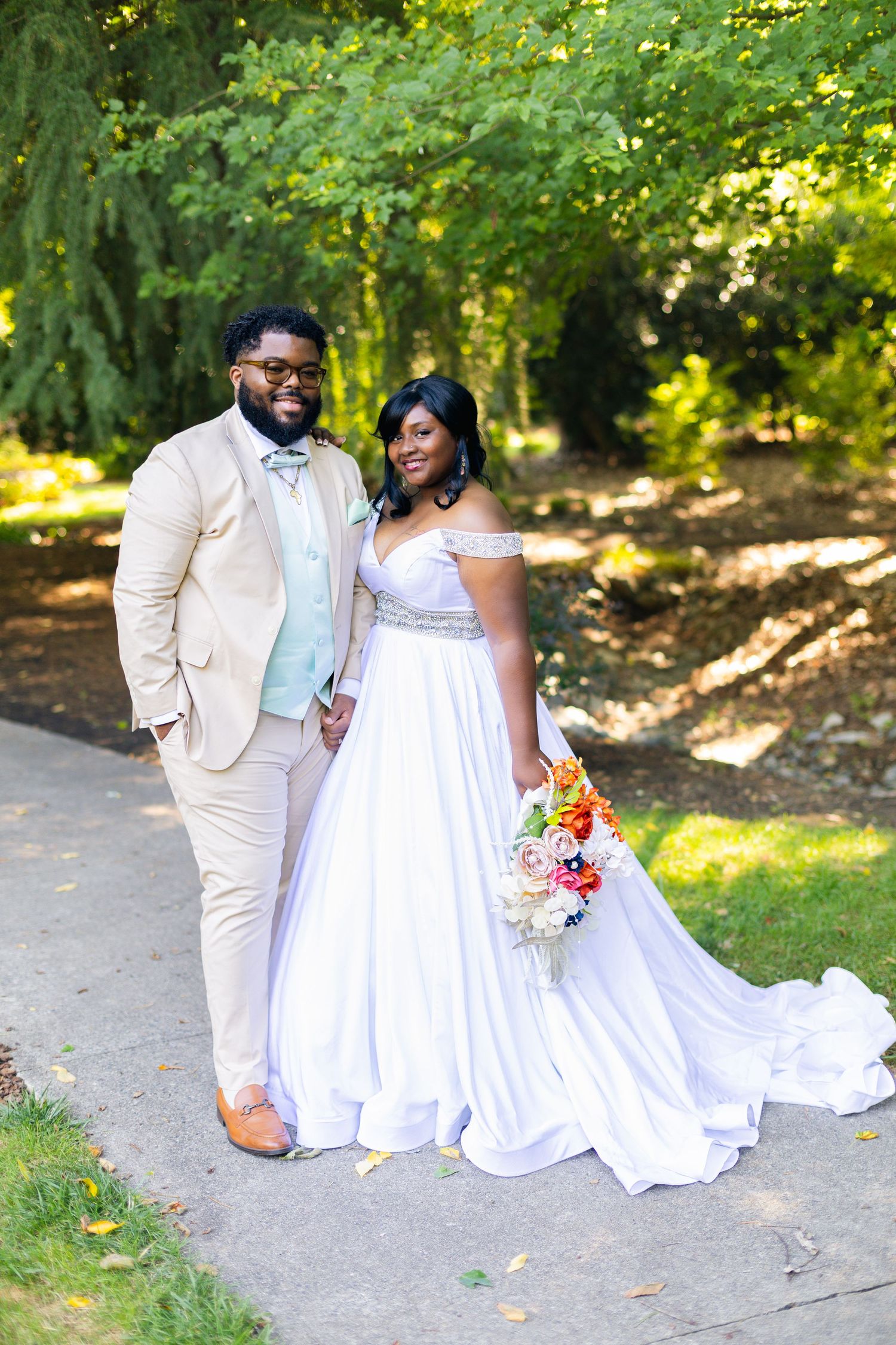 A smiling bride and groom pose outdoors in wedding attire, she holds a colorful bouquet, surrounded by lush green trees.