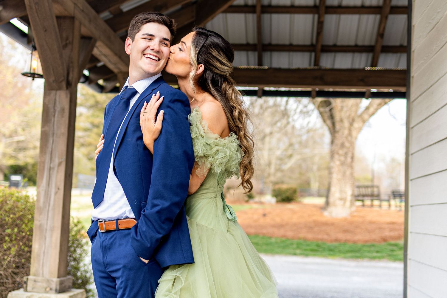 A couple in formal attire embraces outdoors, with the person in blue holding the one in a sage green dress.