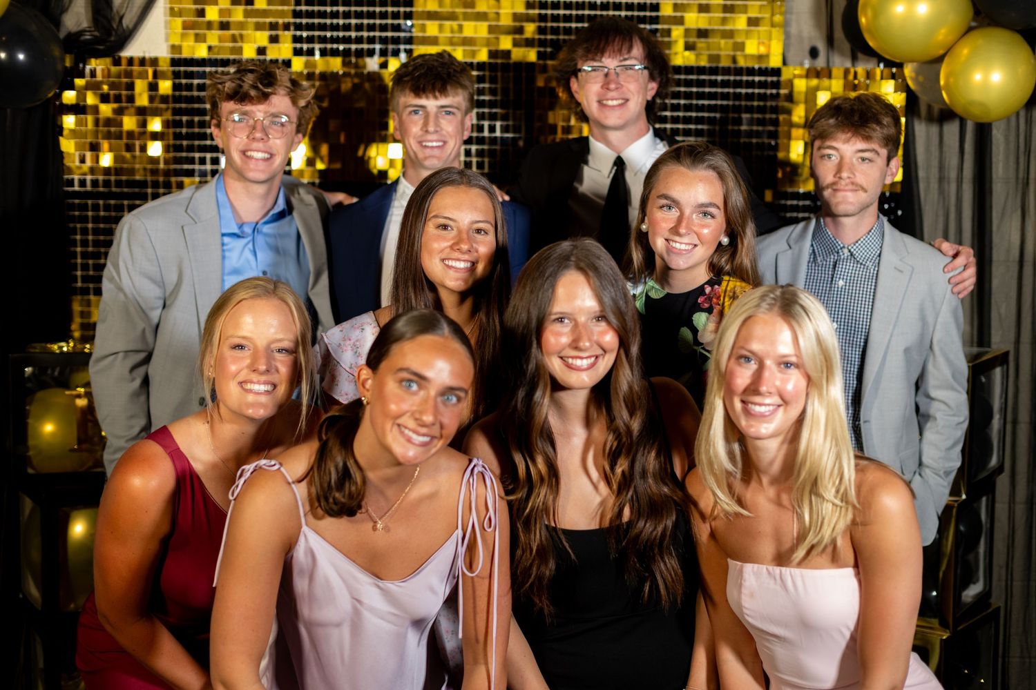 Group of teens in formal attire smiling at prom event with gold balloons and glittery backdrop.
