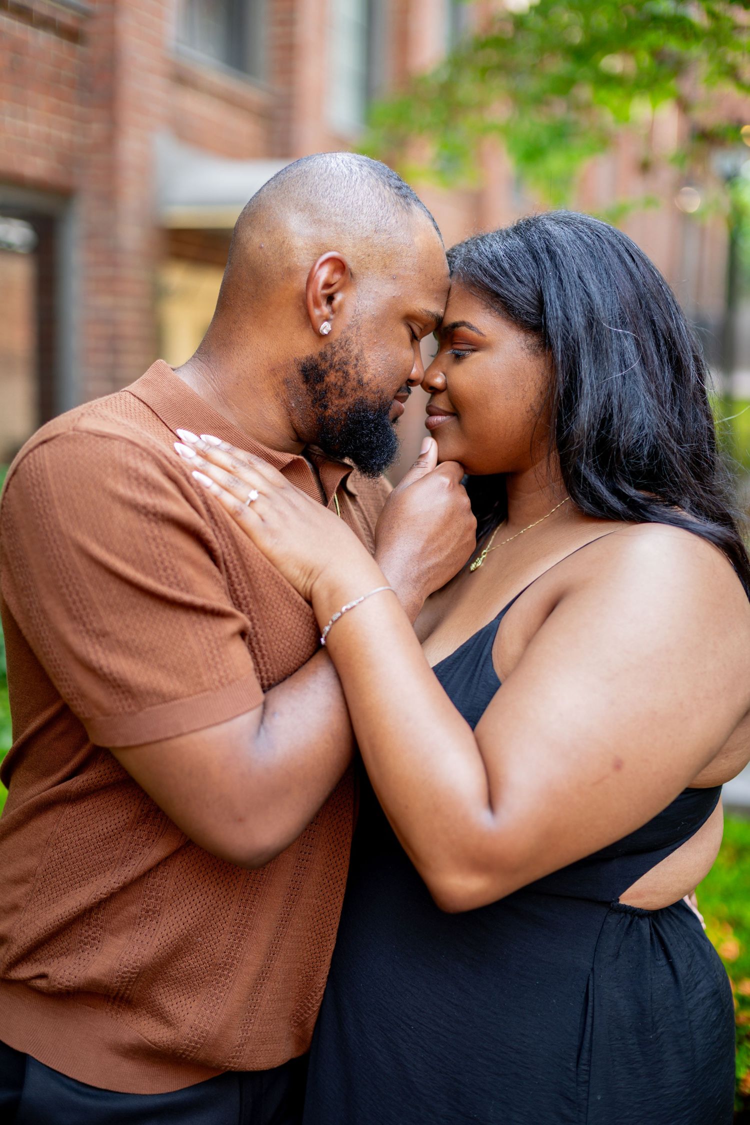 A couple sharing a romantic moment outdoors, touching foreheads and smiling near a brick building.