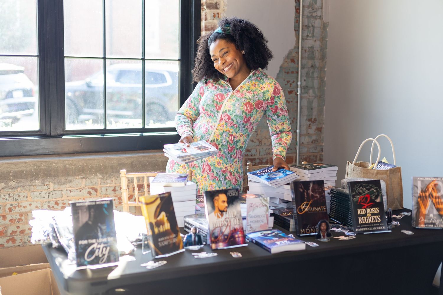 Woman smiling at a book display table with various novels and tote bags at an indoor book fair event.
